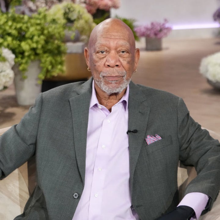 A man sits on a couch with potted plants in the background, wearing a gray blazer and pink shirt, looking at the camera