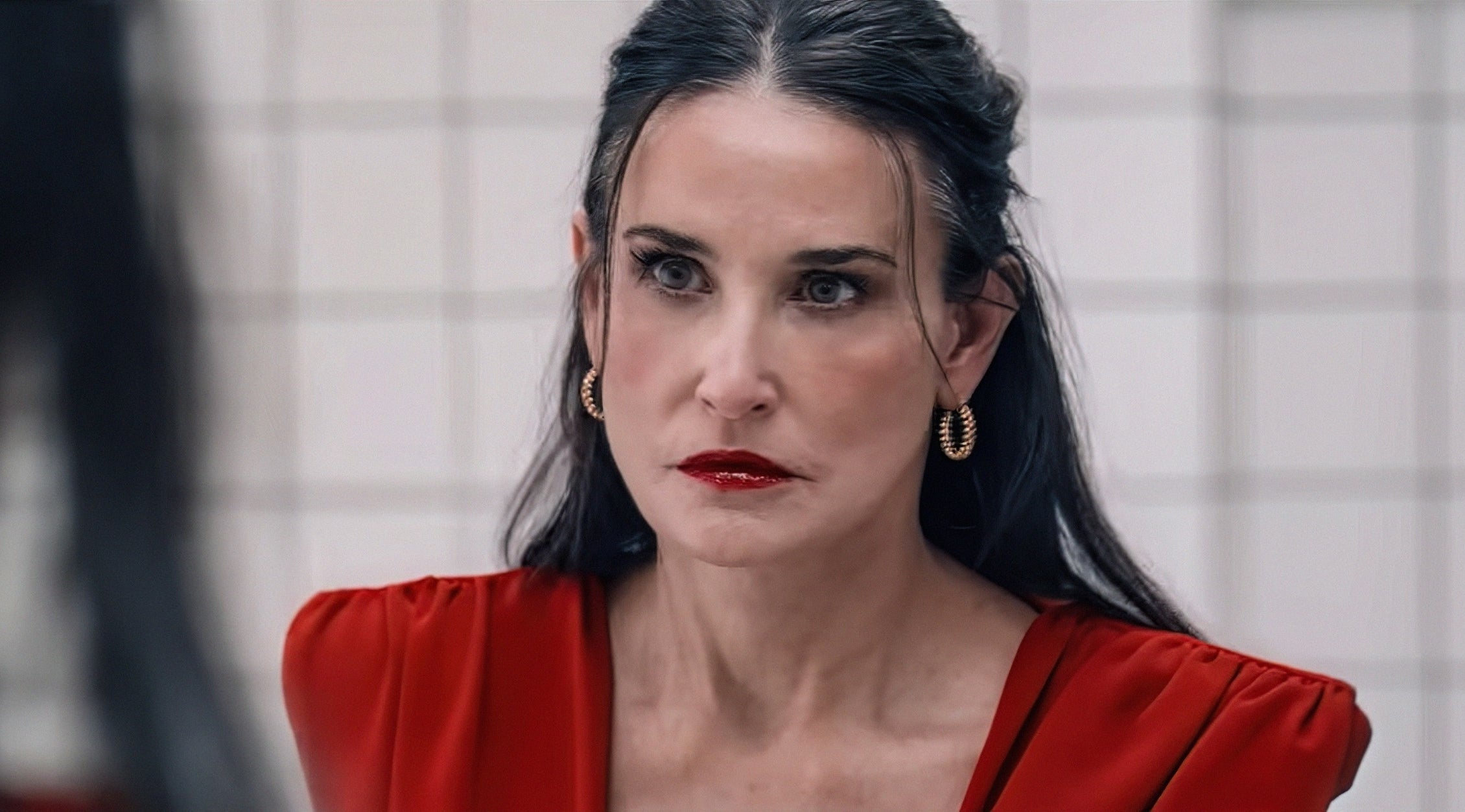 Demi Moore, with long dark hair and gold hoop earrings, appears serious, wearing a stylish V-neck top, gazing into a mirror in a tiled room. This is a scene from &quot;The Substance&quot;