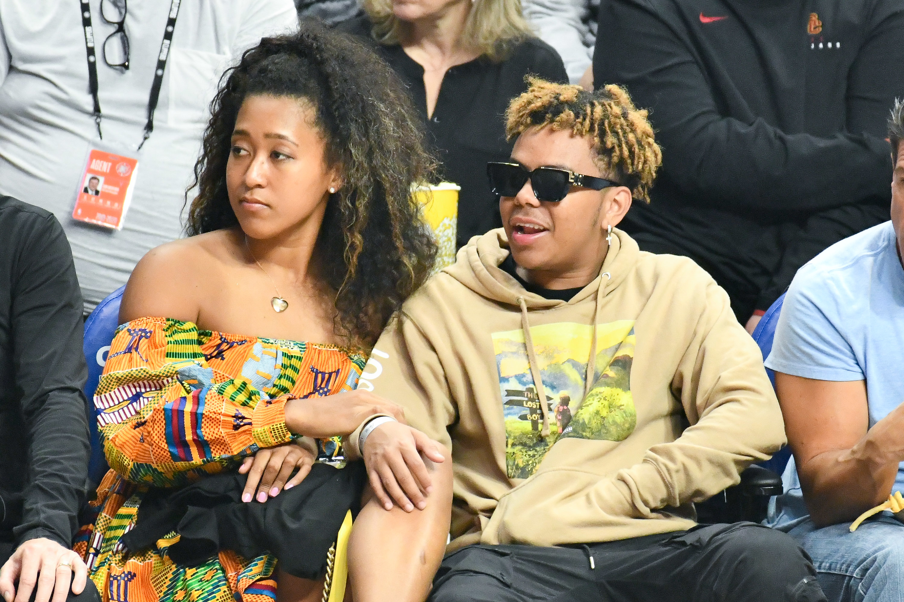 Naomi Osaka and Cordae sitting courtside at a basketball game. One wears a colorful patterned dress, the other wears a hoodie and sunglasses