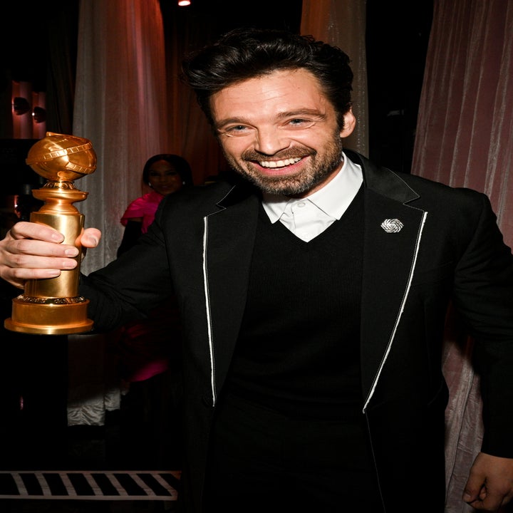 Sebastian Stan smiling and holding a trophy in a suit with a white shirt and black sweater at an award event