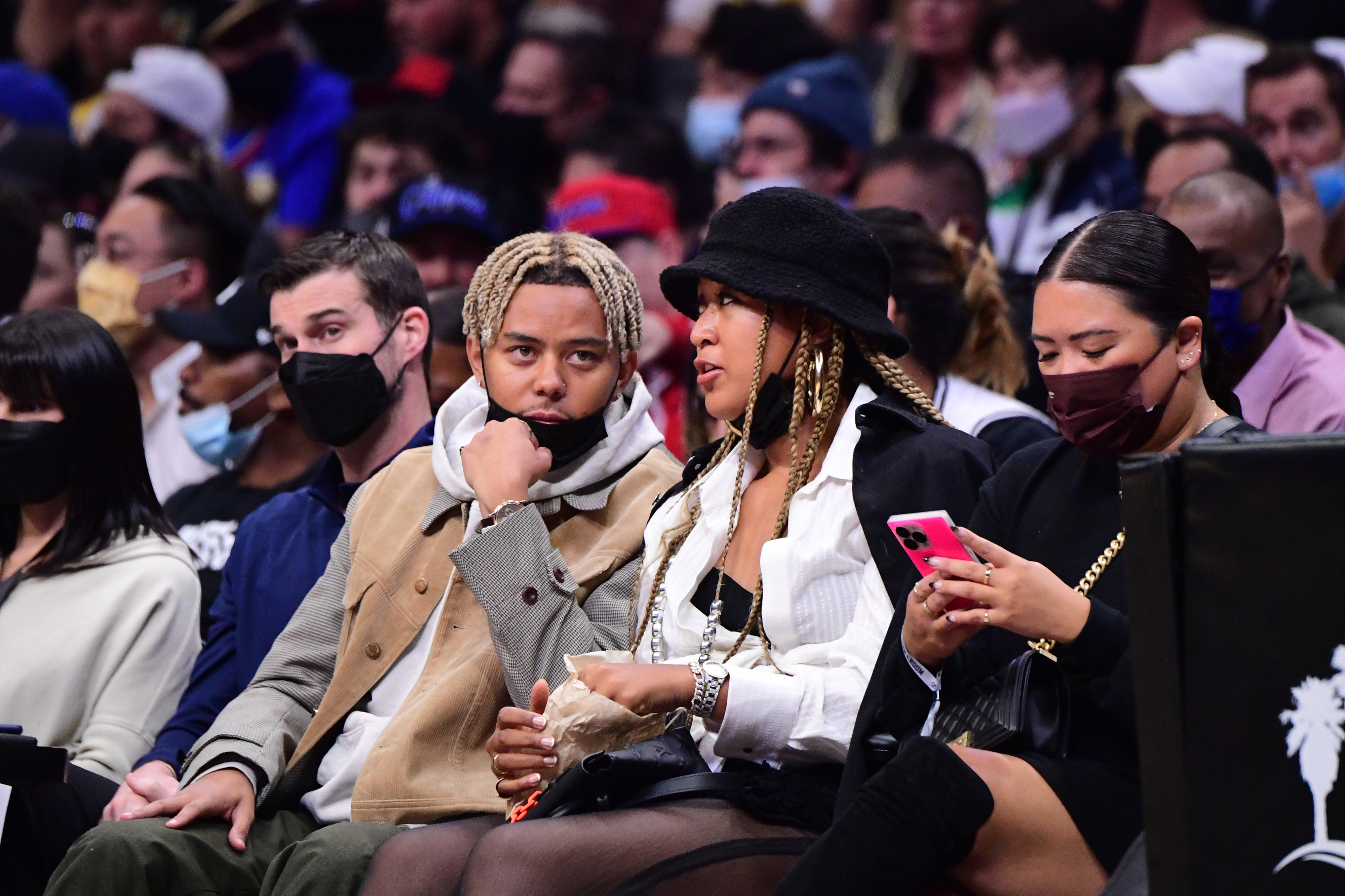 Cordae and Naomi Osaka sit courtside at a sports event, engaged in conversation. The woman wears a black hat and coat, while the man wears a tan jacket