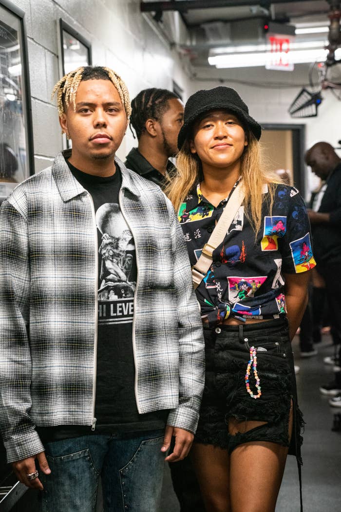 Cordae and Naomi Osaka pose in a hallway. Cordae wears a patterned shirt and shorts. Naomi wears a plaid jacket and tee. People and decor are in the background