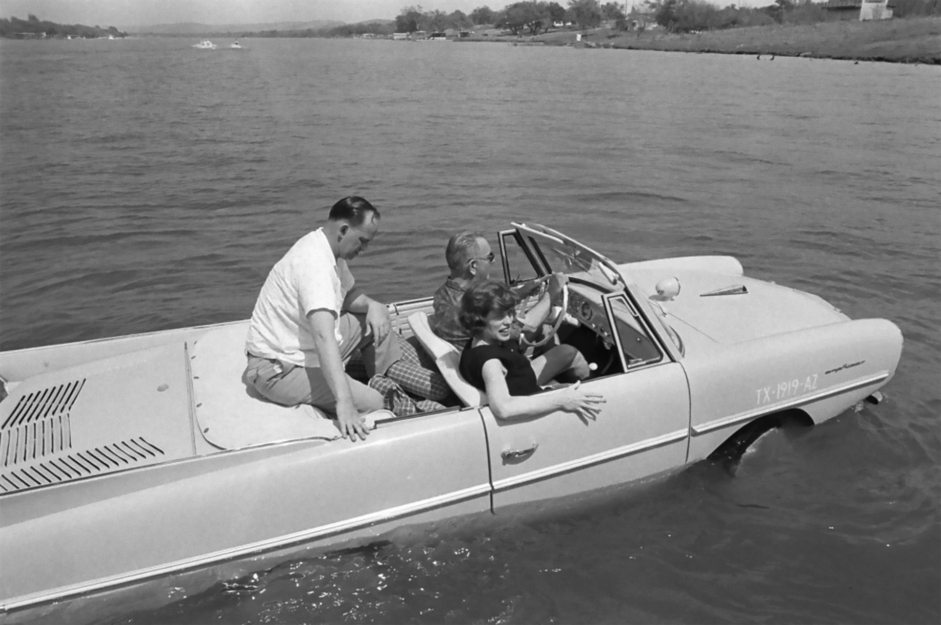 A vintage car is floating on water with three people inside, two men and a woman, enjoying a ride near the shore