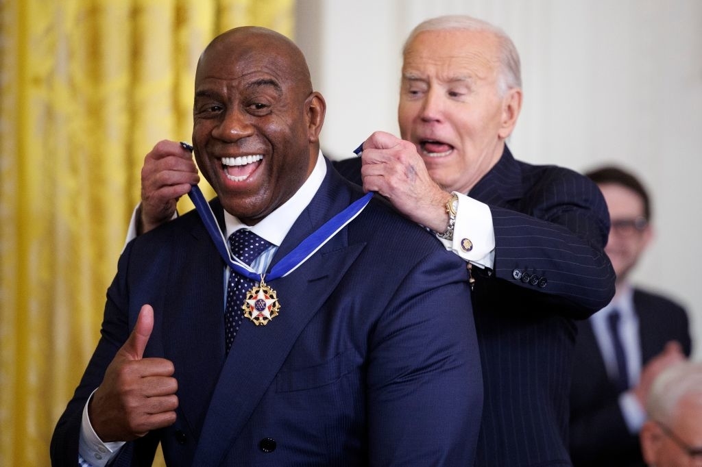 A smiling man receives a medal from another man during a formal ceremony. The recipient wears a dark suit and gives a thumbs-up