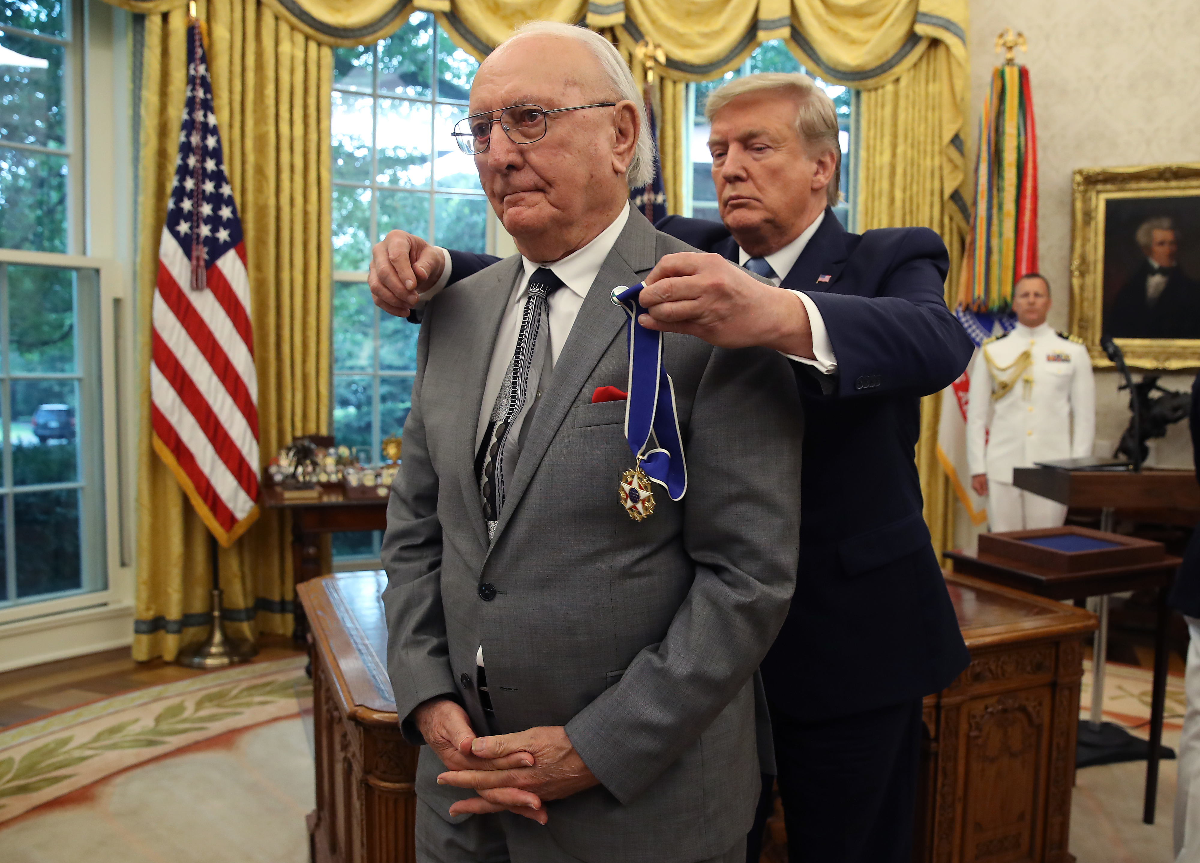 An elderly man stands receiving a medal from a man in a suit in an official room with flags and a large window in the background