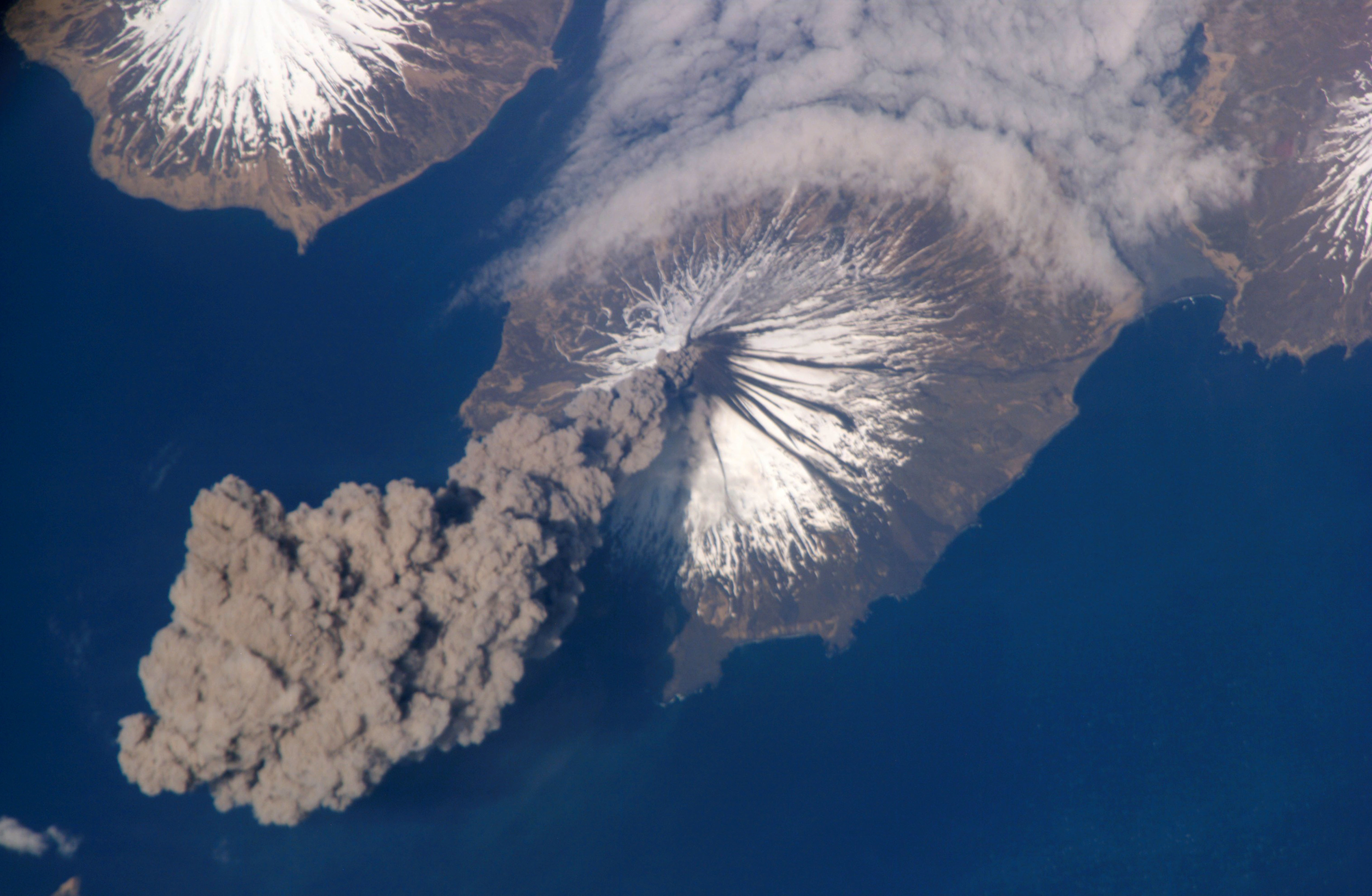 Aerial view of a volcanic eruption with ash plume, snow-capped peaks, and surrounding ocean