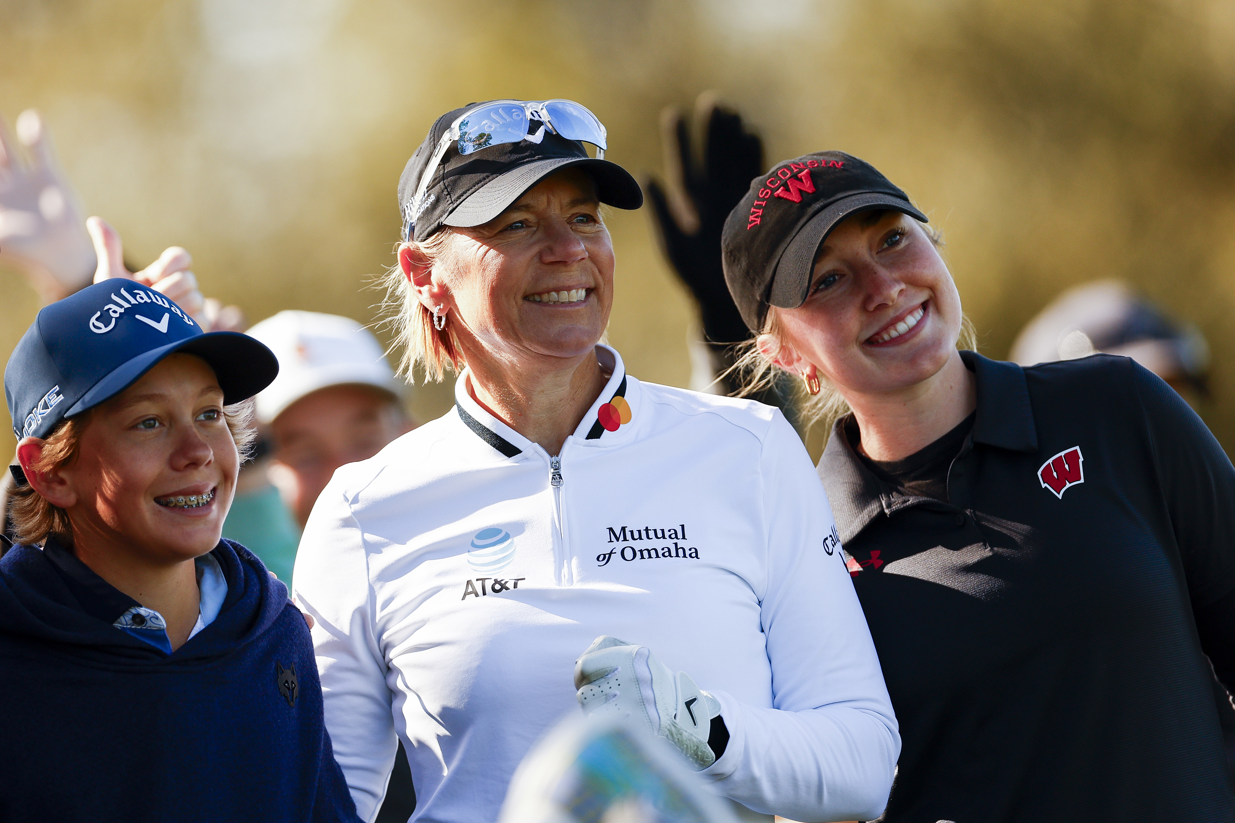 Three people smiling at a golf event, with one wearing a golf cap and sporty jacket