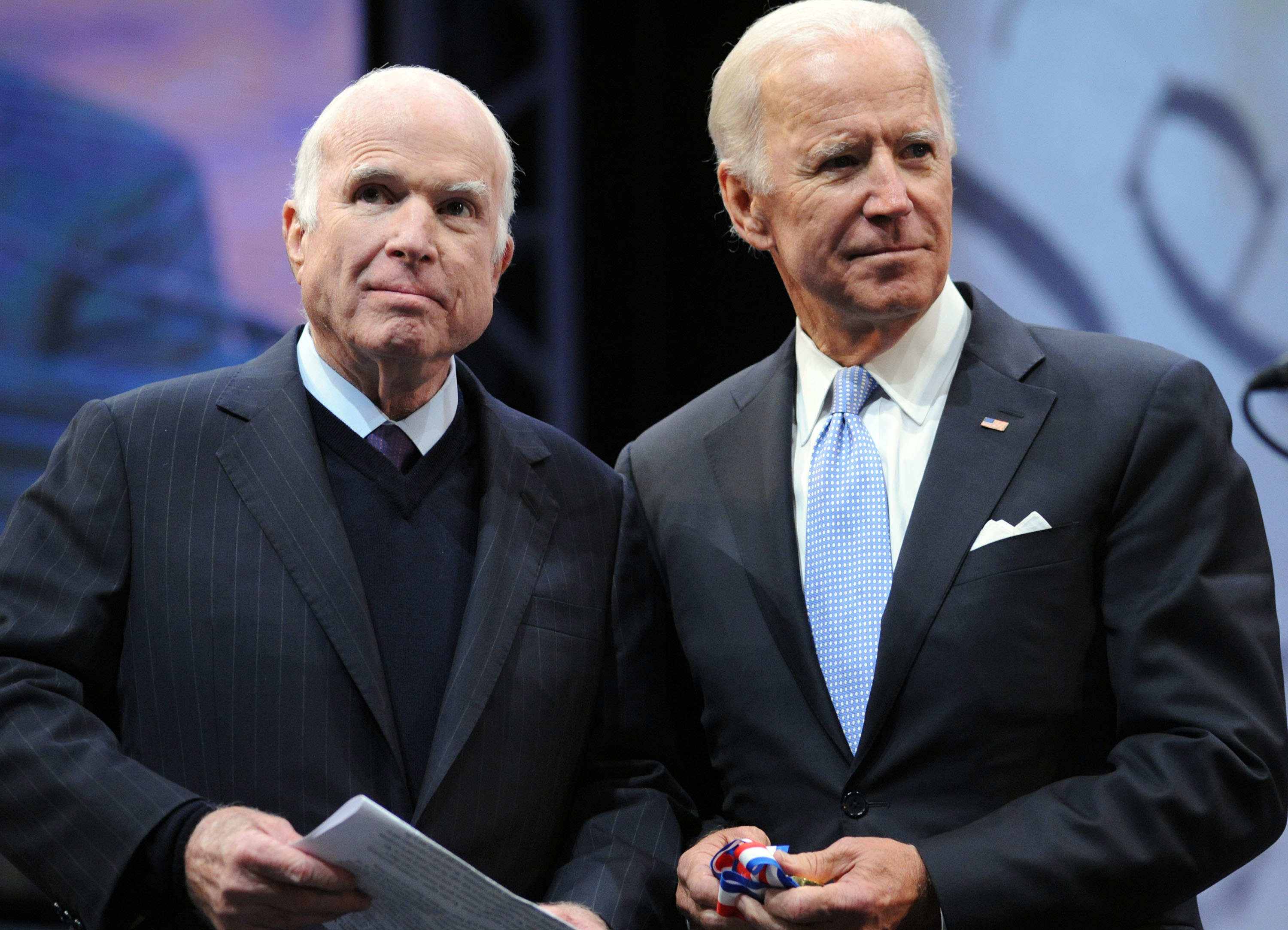 Two men in suits stand together at an event, holding papers and looking thoughtfully ahead