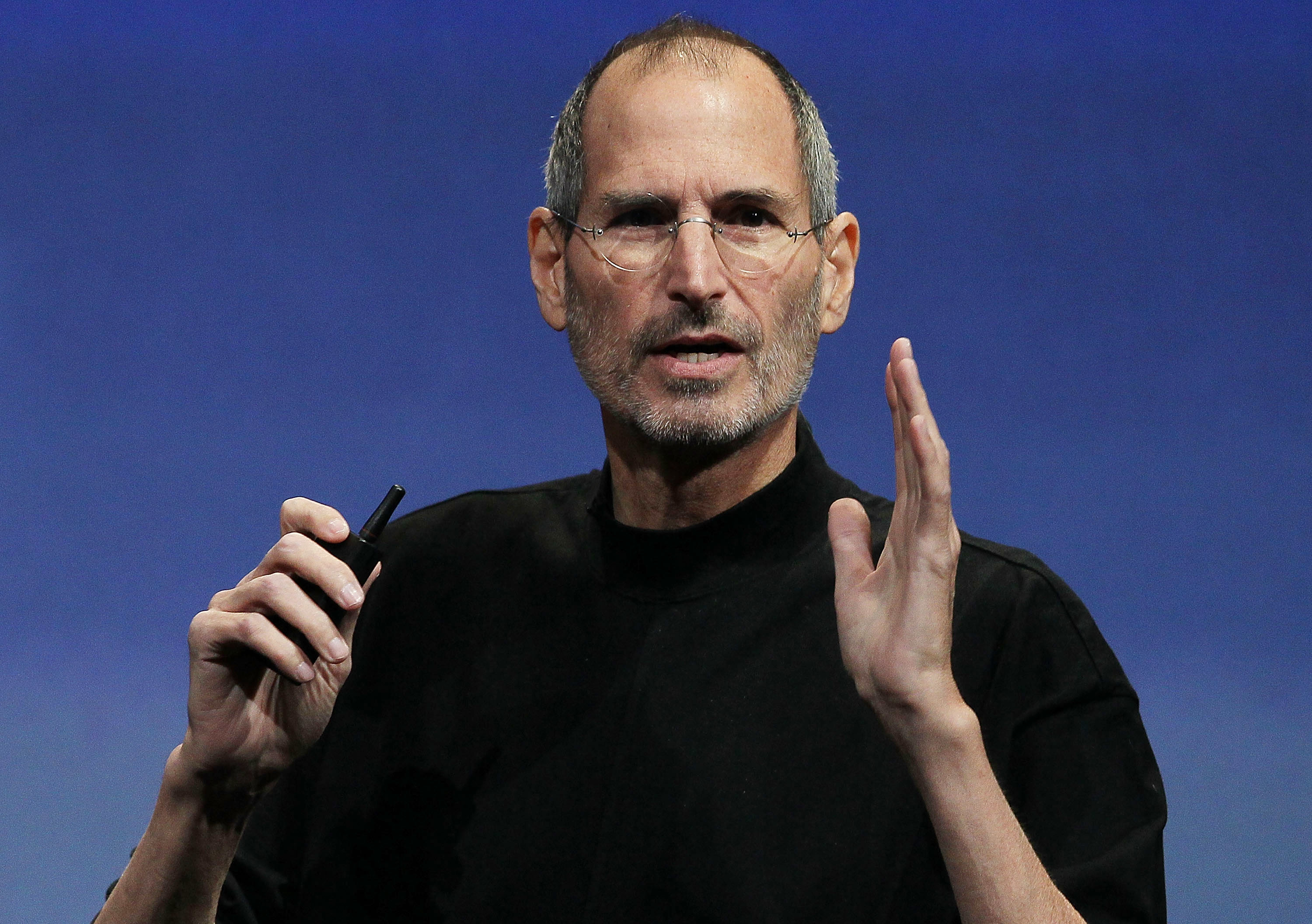 A person speaking on stage, dressed in a black turtleneck, holding a small device, gesturing with one hand