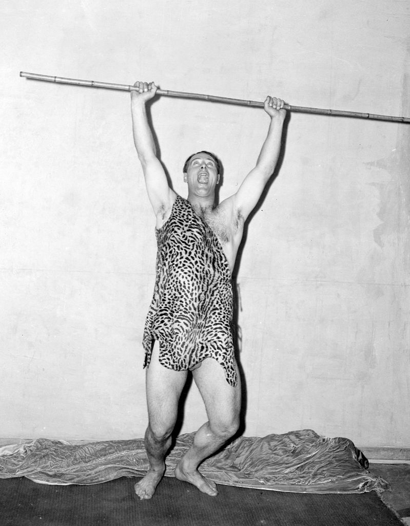 Man in leopard print toga poses with a spear overhead, striking a triumphant pose