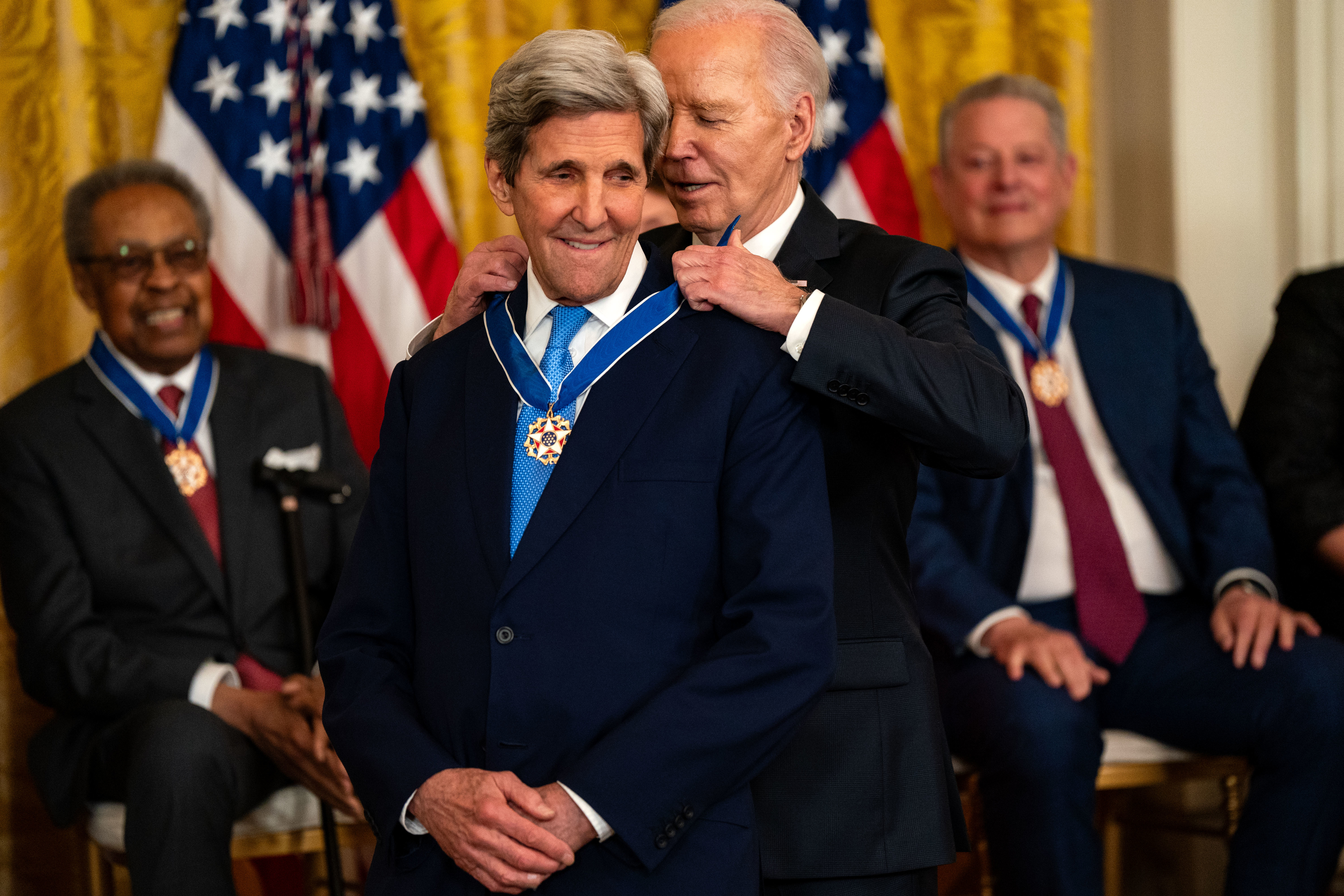 A man awards another man a medal as two seated men watch in a formal ceremony setting