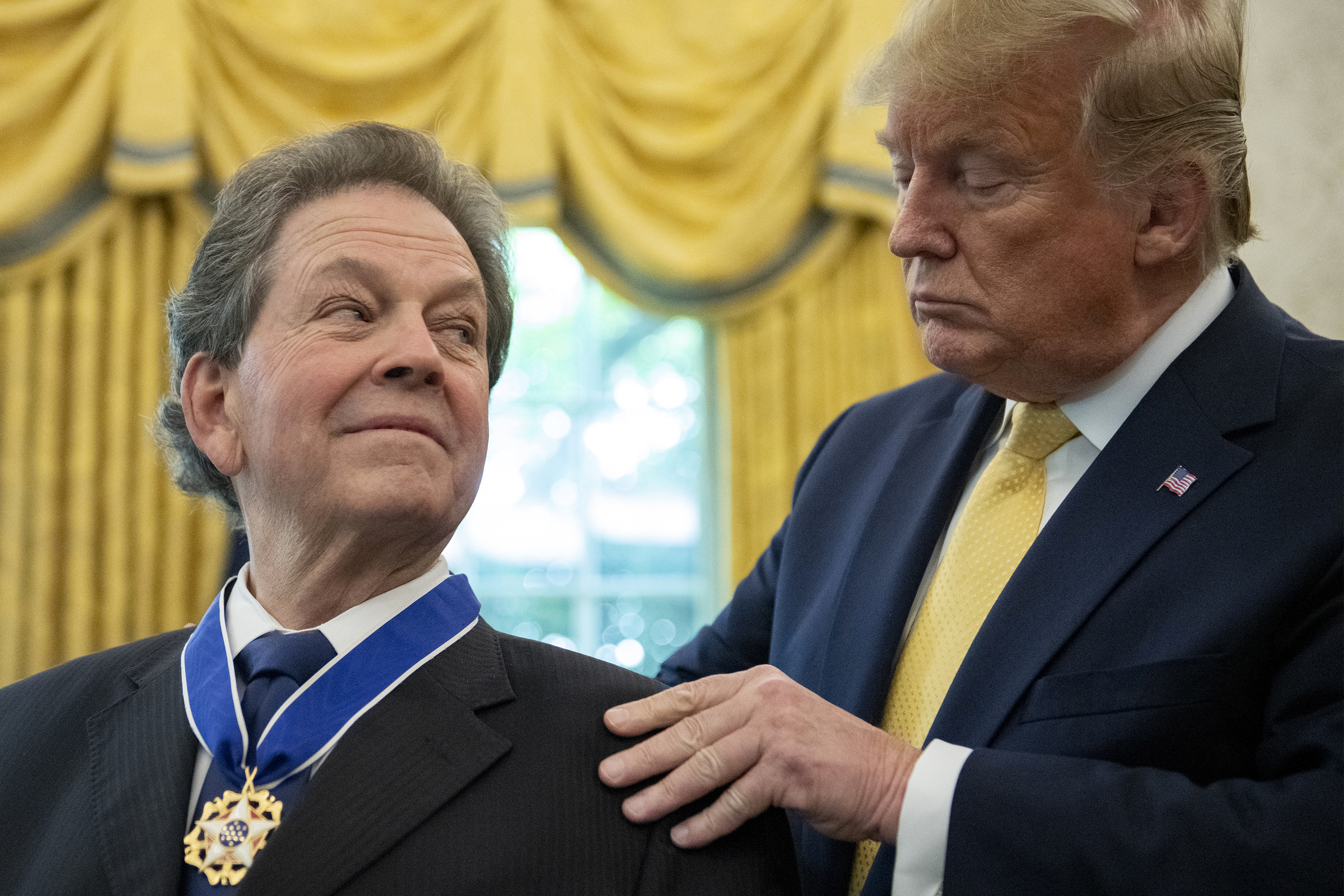 A person is awarding a man a medal in a formal ceremony, both in suits, with gold curtains in the background