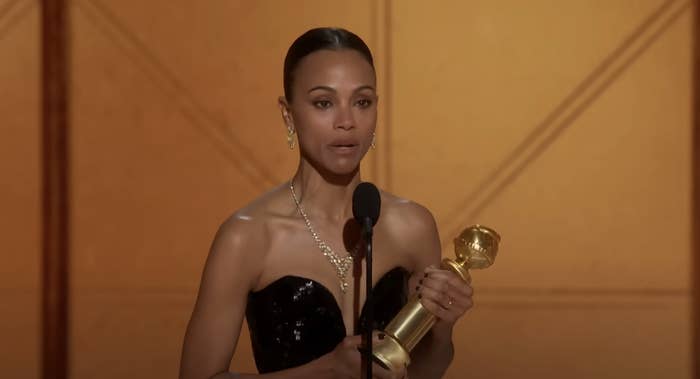Zoe Saldaña on stage in a strapless dress holding an award and speaking into a microphone
