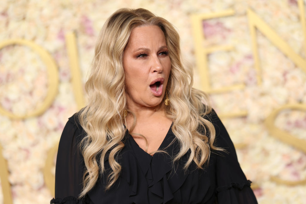 Jennifer with wavy hair wearing a black ruffled top, making an expressive face against a floral backdrop at a public event