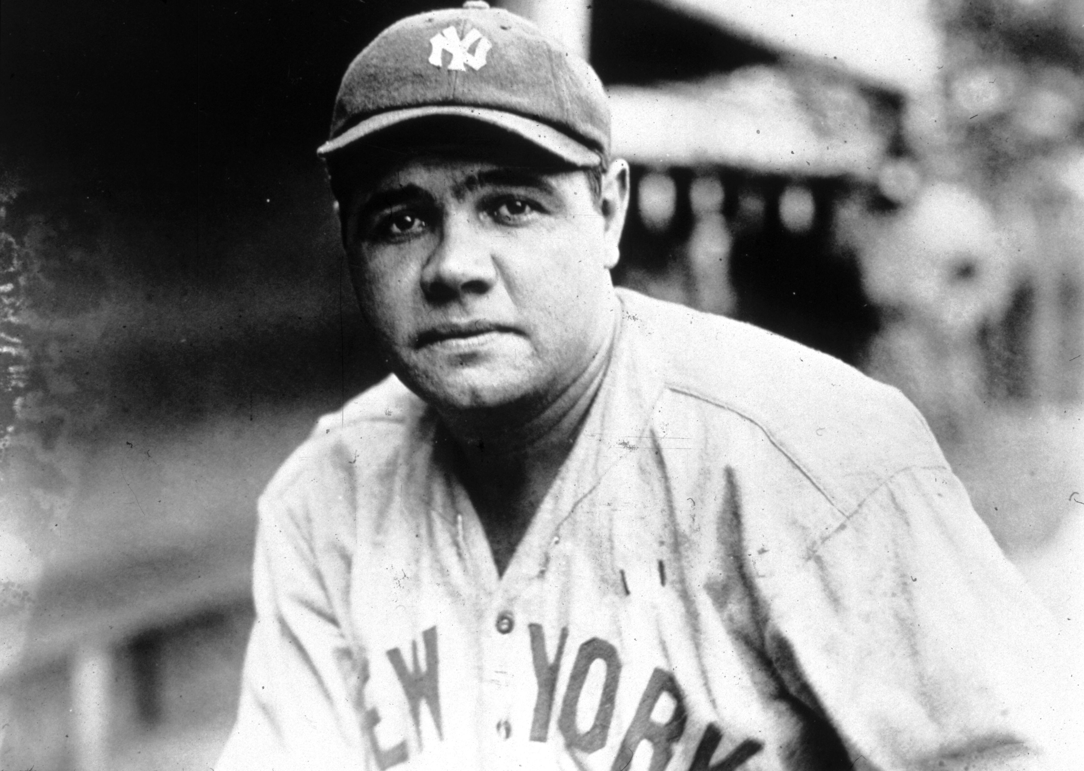 A baseball player in a vintage Yankees uniform and cap looks at the camera. The setting indicates an early 20th-century baseball environment