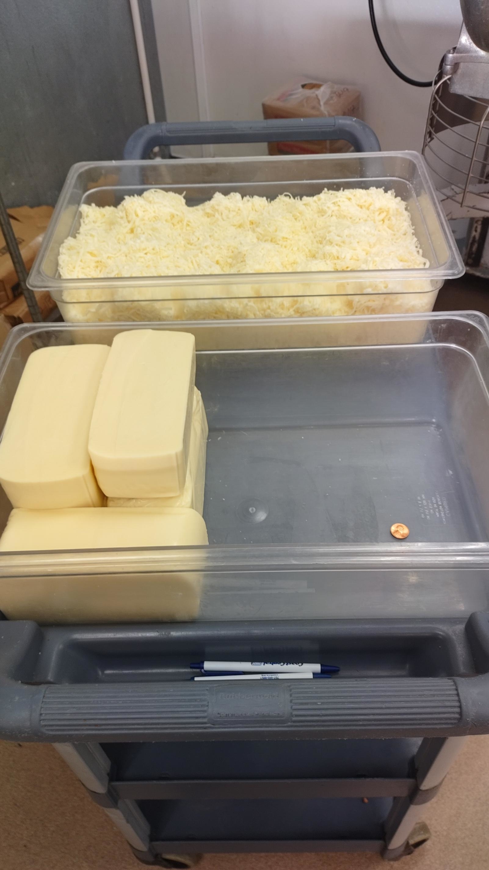 A cart with a tray of shredded cheese, a tray of cheese blocks, a single penny, and a pen