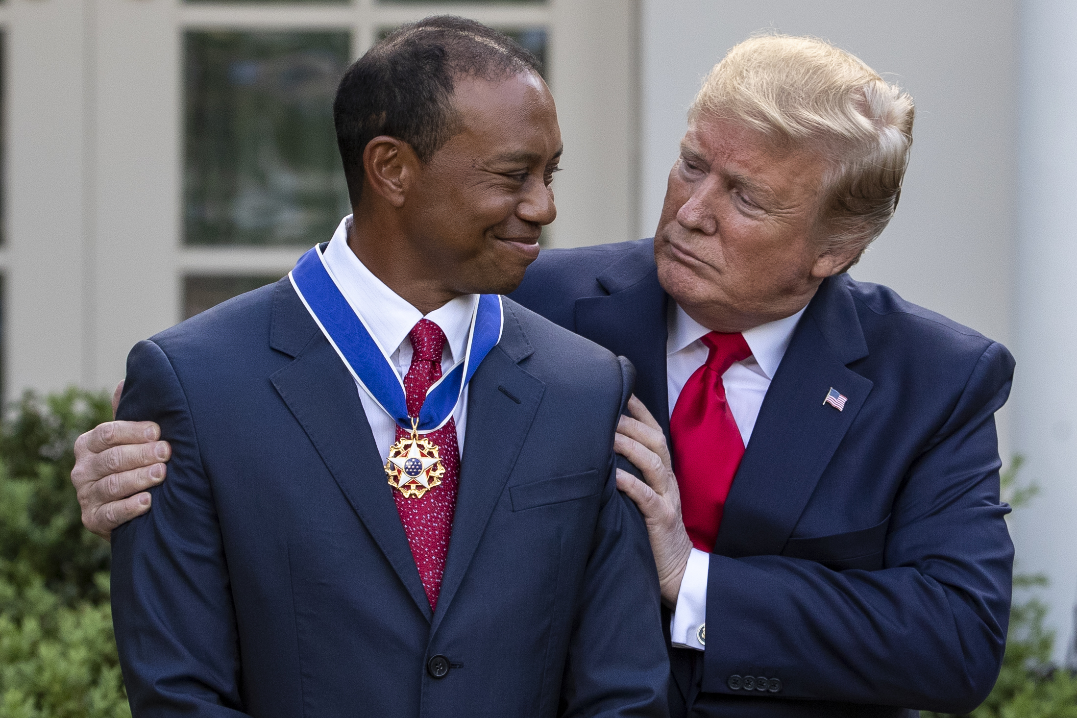 Two men in formal suits; one wearing a Medal of Freedom. The other places his hands on his shoulders in an outdoor ceremony setting