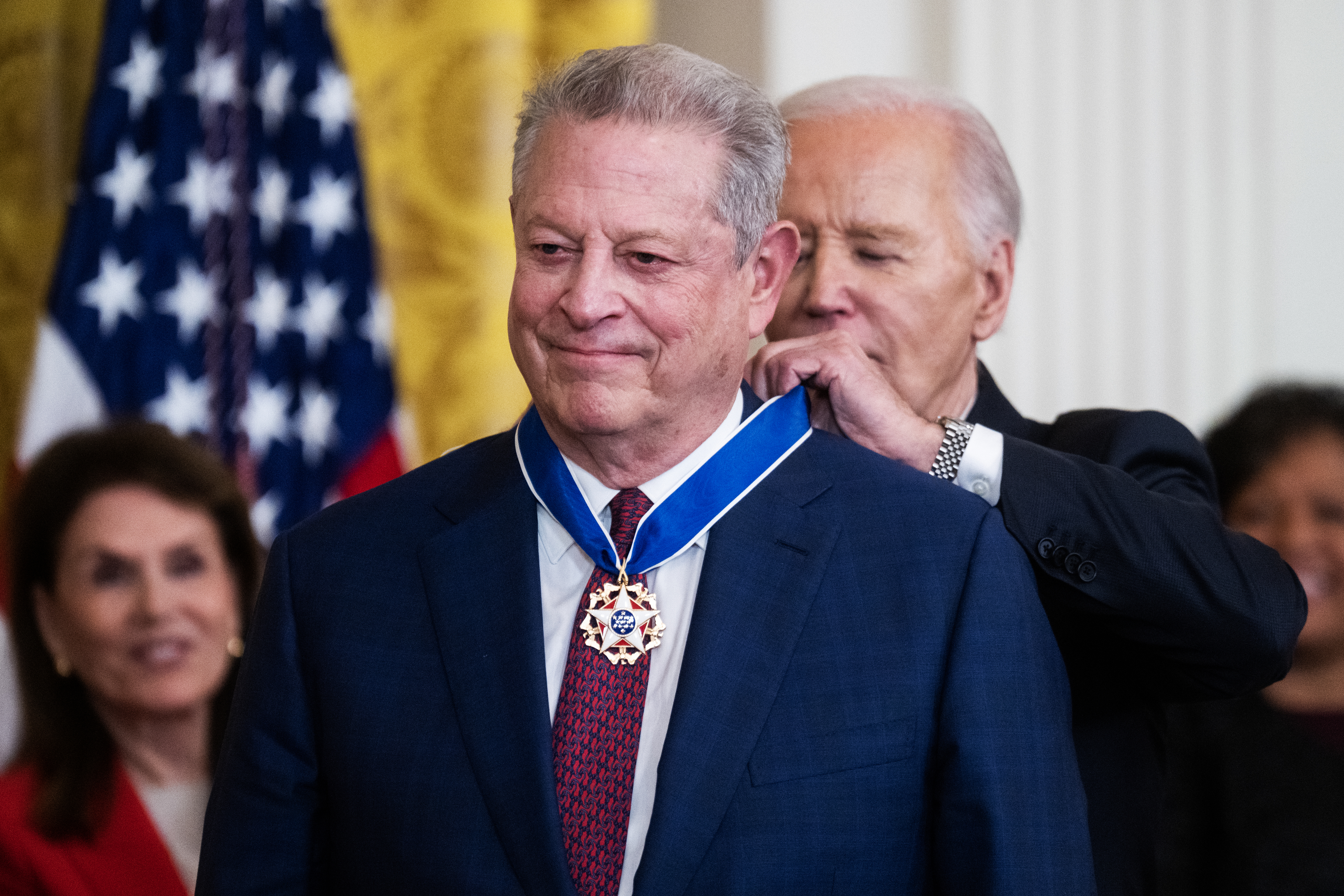 Person receiving a medal from another person at a formal event with the U.S. flag visible in the background