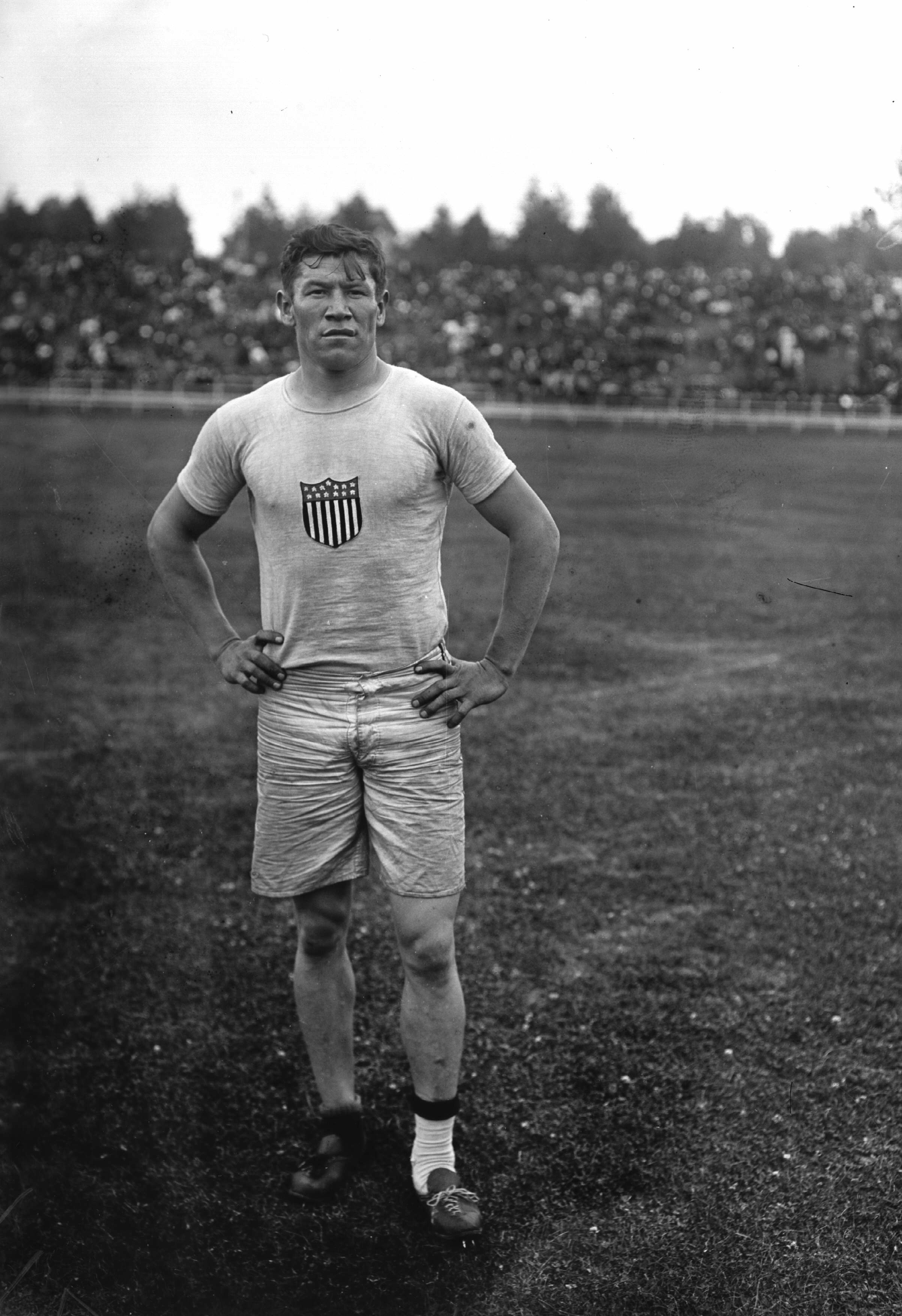 Athlete stands on a field, hands on hips, wearing a shirt with a shield emblem and shorts; crowd in background. Early 20th-century style