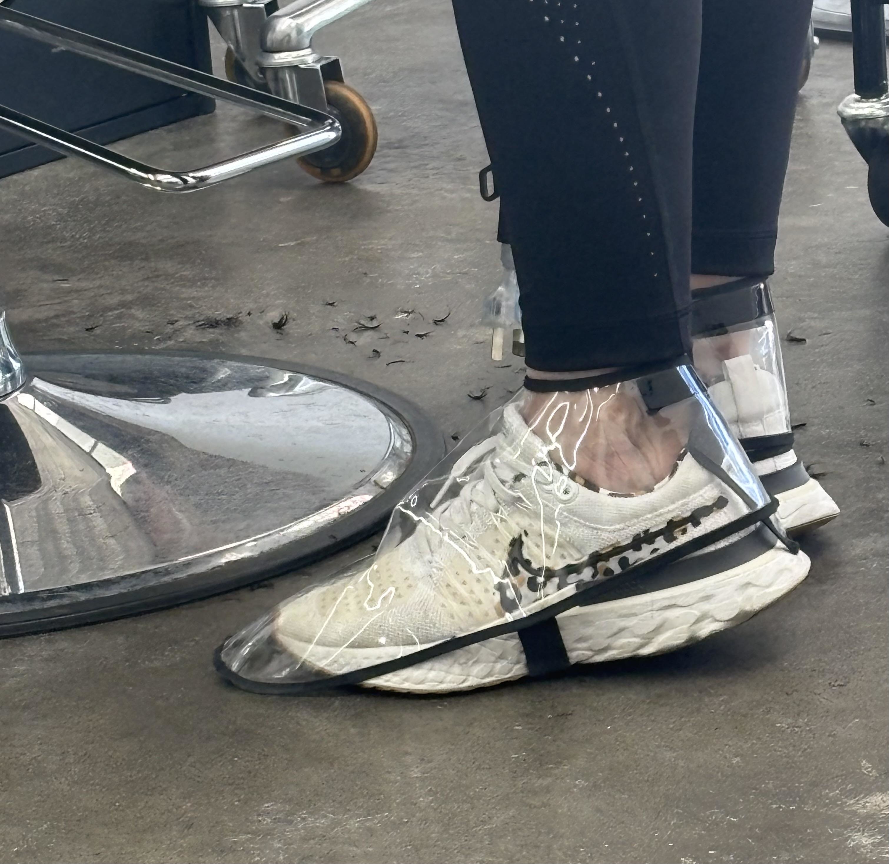 Sneakers are covered in transparent plastic shoe covers, worn by a person seated on a hair salon chair, with hair clippings on the floor