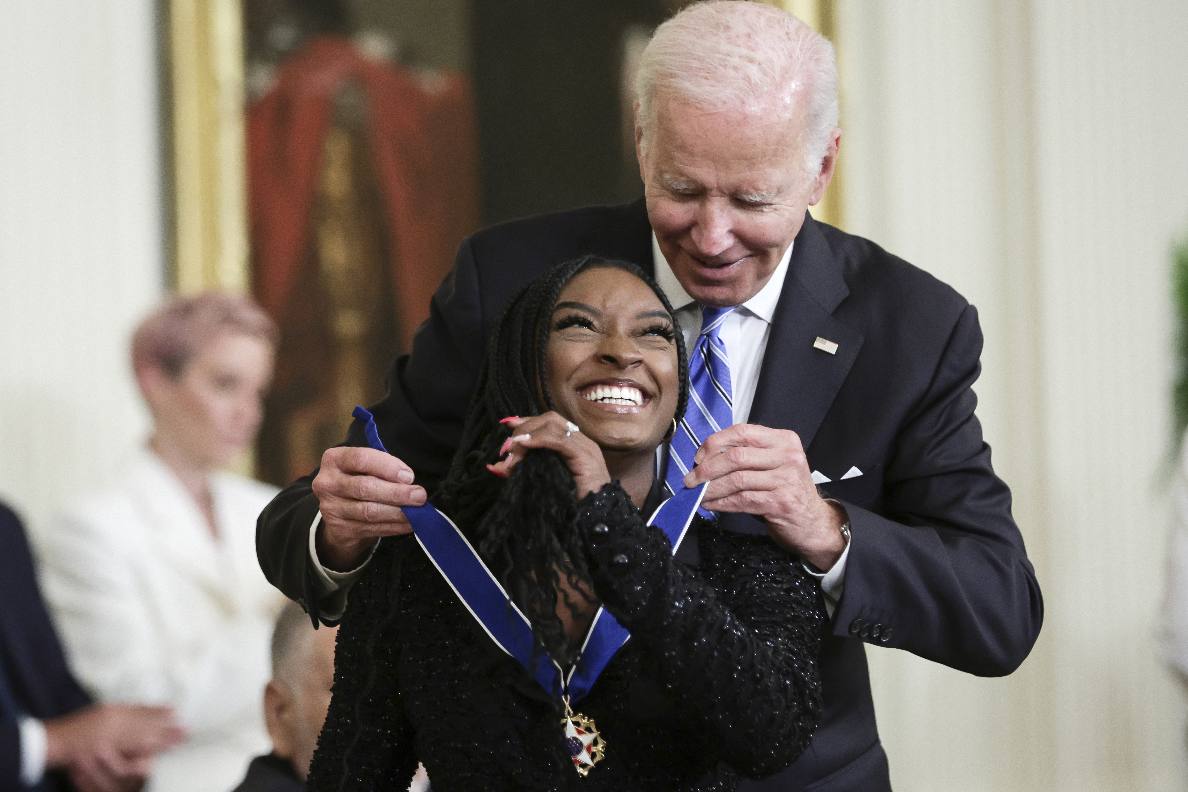 Person receiving a medal from another person in a formal setting, both smiling and embracing