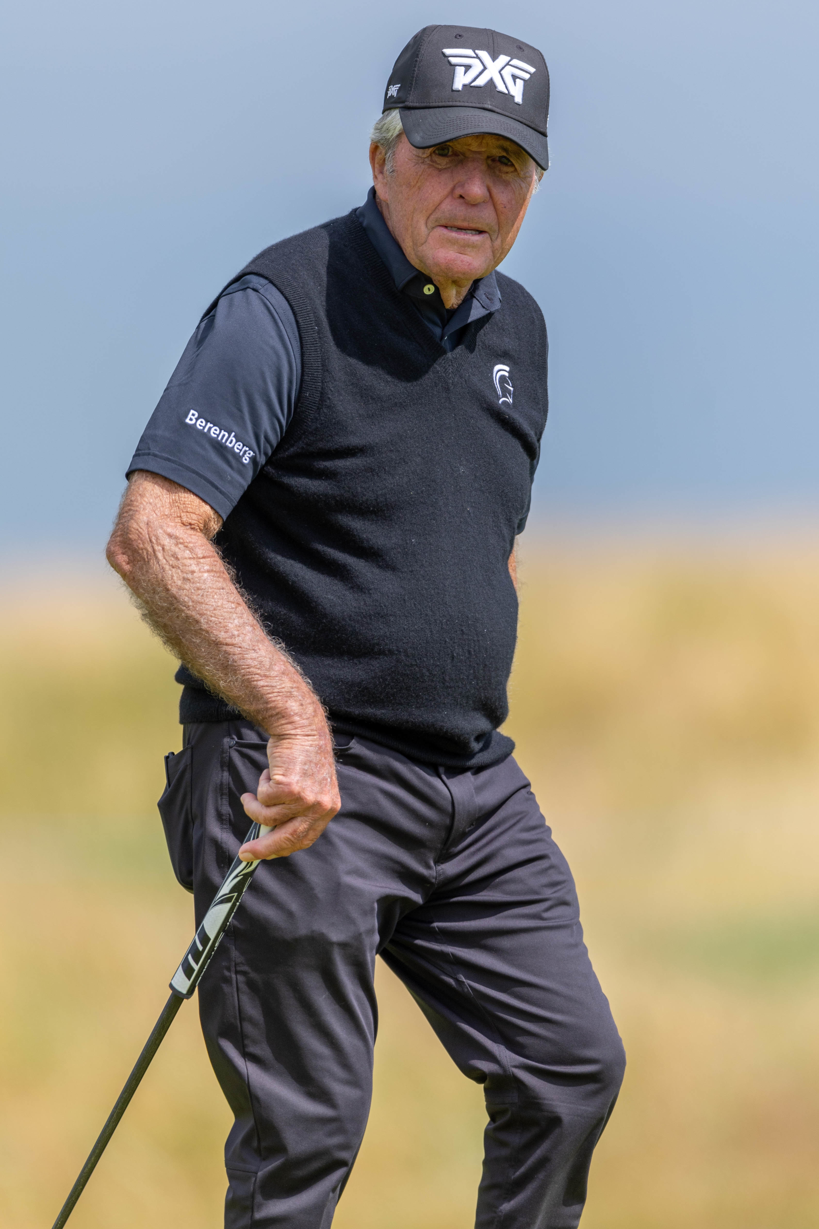 An older adult golfer on a golf course holds a club, wearing a sporty outfit with a cap