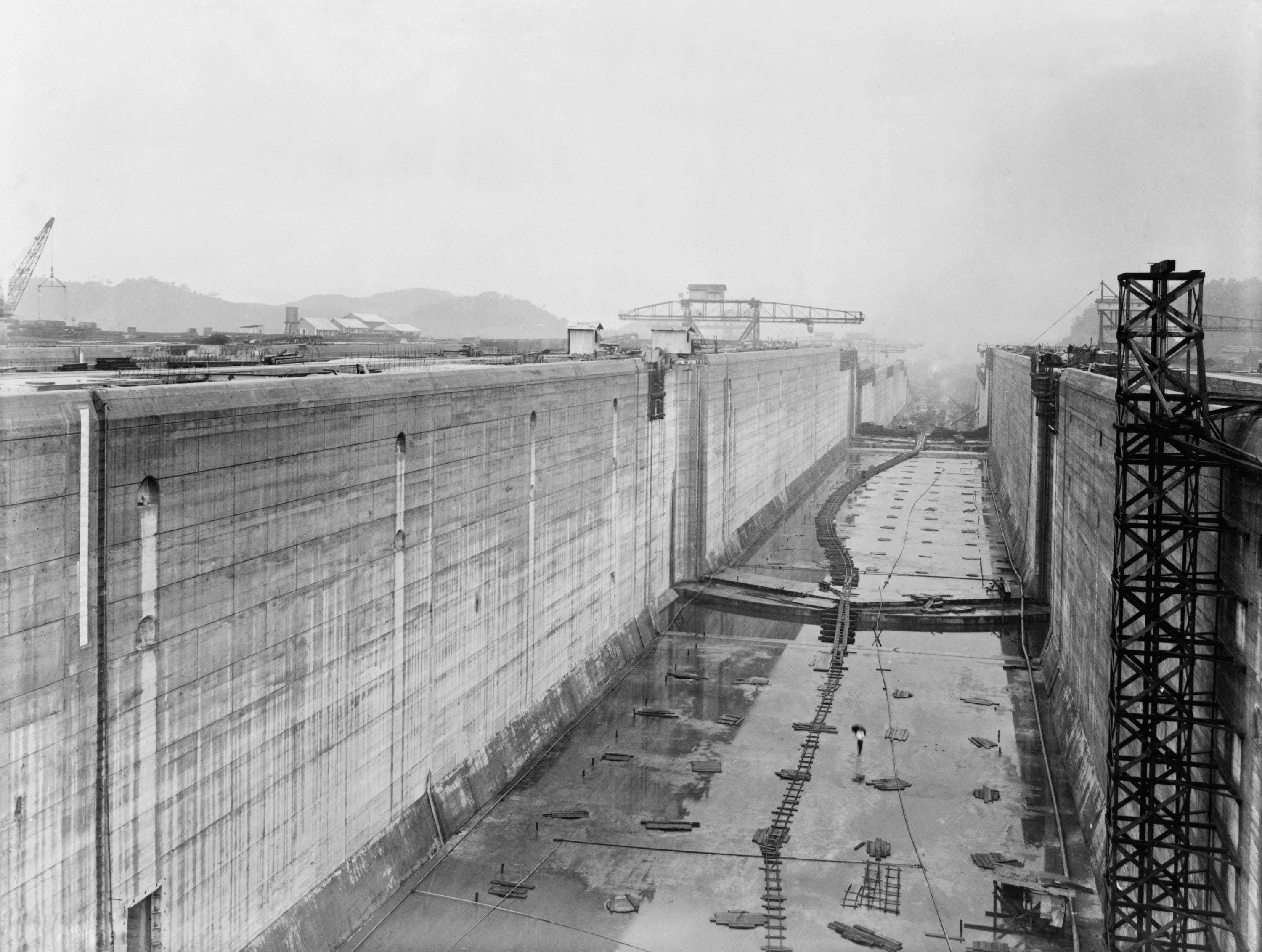 Historic black-and-white photo of the Panama Canal under construction, featuring partially built massive concrete walls and large cranes in the distance