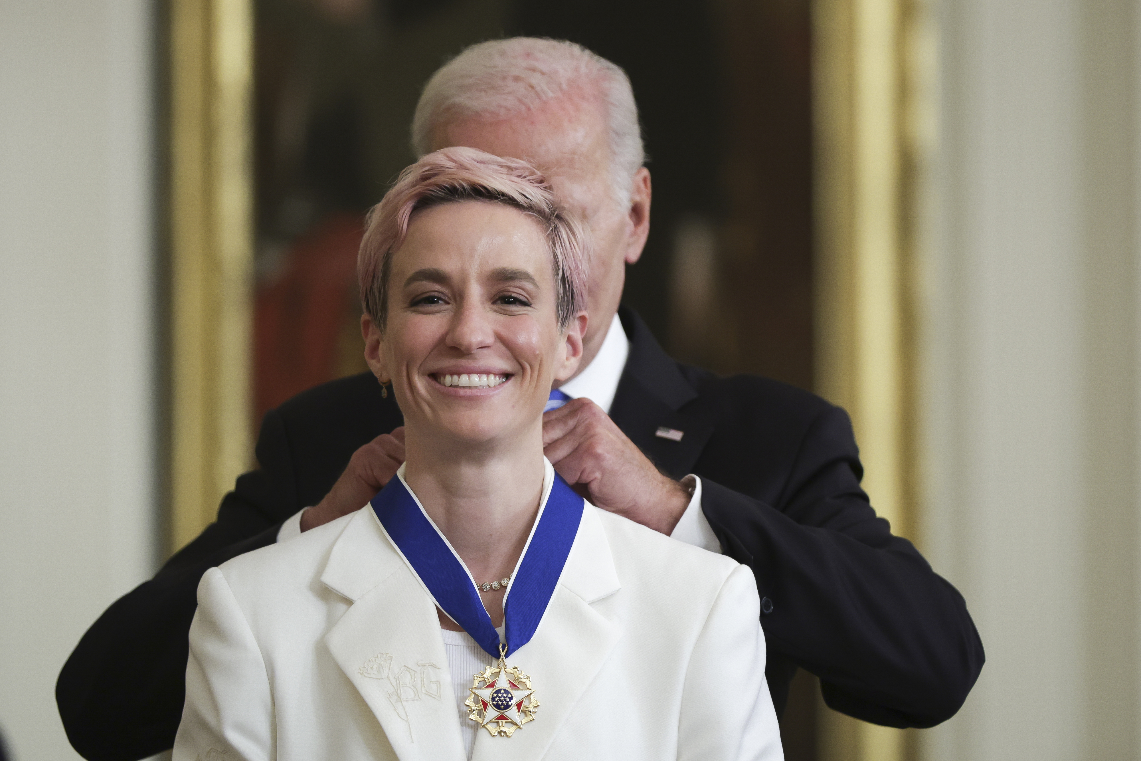 Person in a white suit receiving a medal with a ribbon from another individual standing behind them