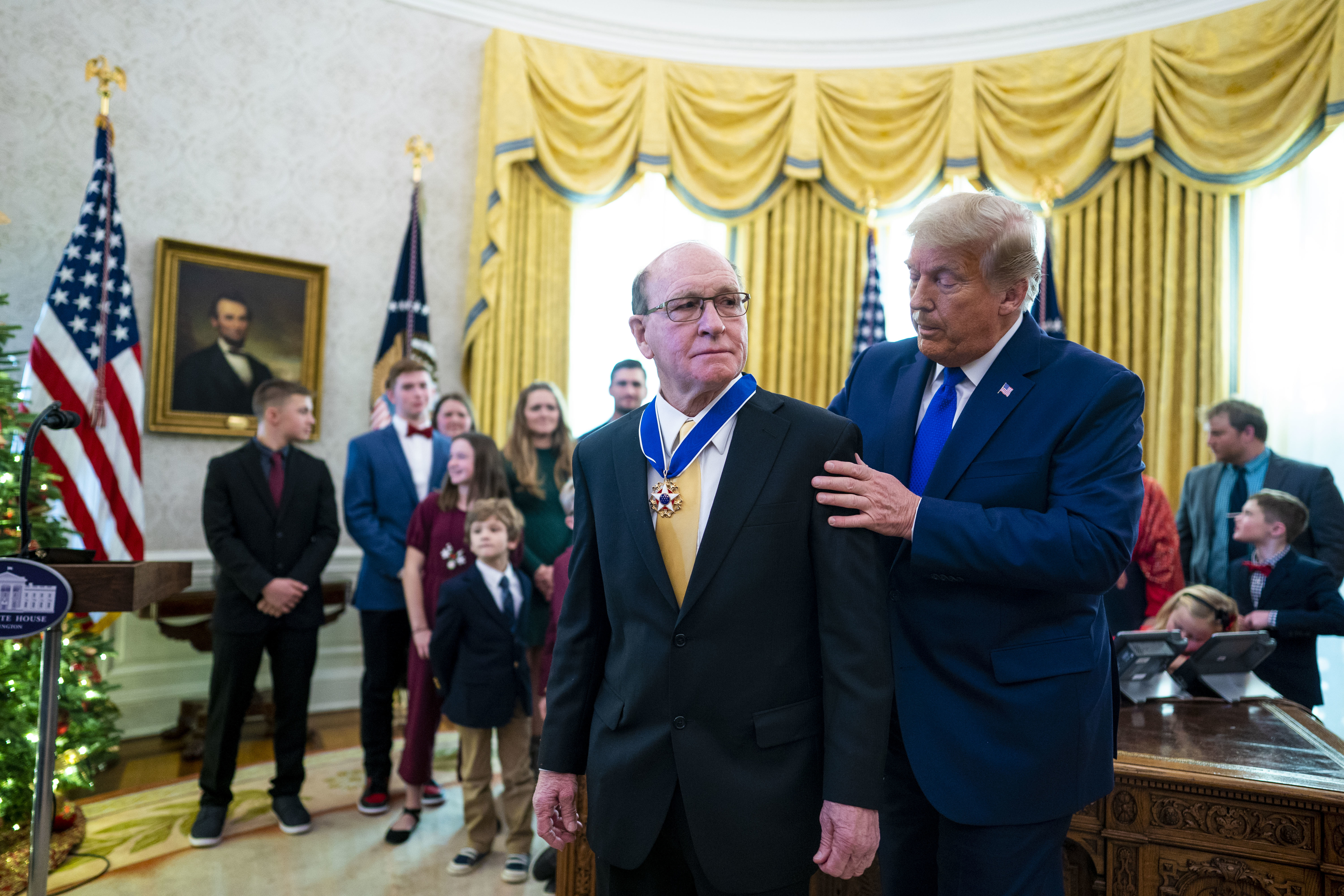 A man receives a medal from a prominent figure in a formal ceremony setting, surrounded by a group of onlookers