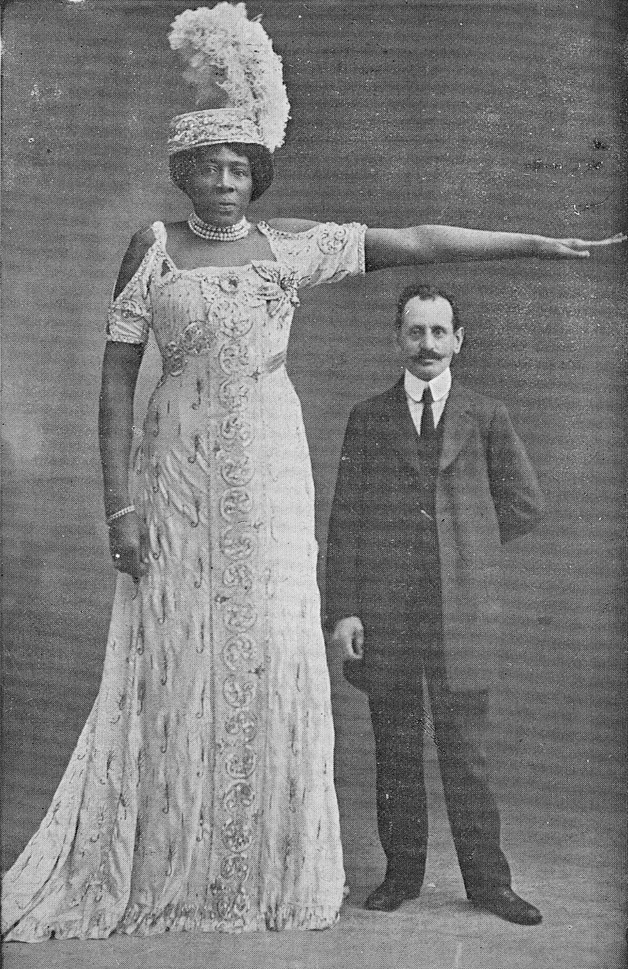 A woman in an ornate, floor-length gown with a feathered headpiece stands beside a man in a suit, showcasing a significant height difference