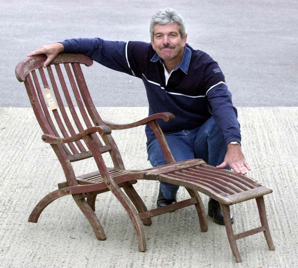 Man smiling and posing with a wooden deck chair in a casual setting