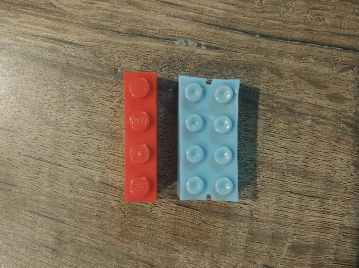 Two rectangular building blocks, one red, one blue, with raised dots, placed on a wood-textured surface