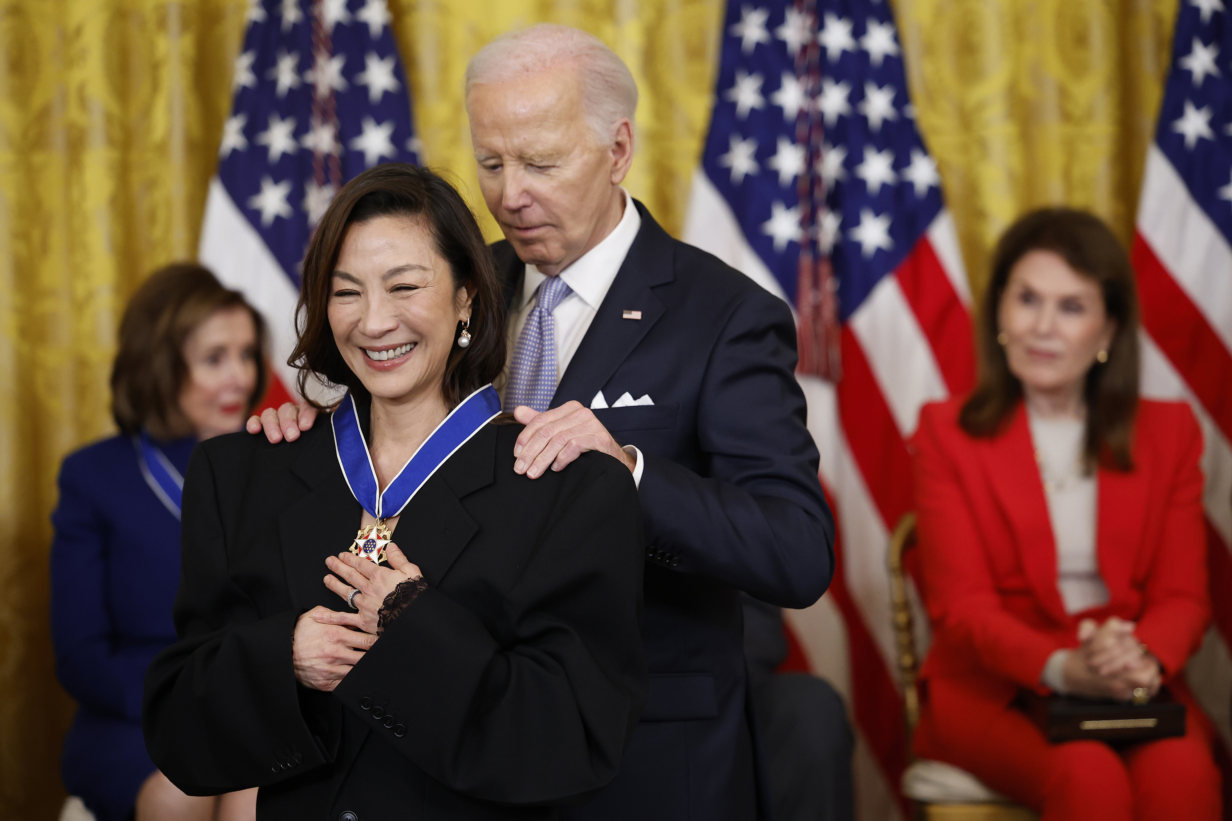 Person receiving an award from a man in a suit, with an audience in the background. Flags are visible in the setting