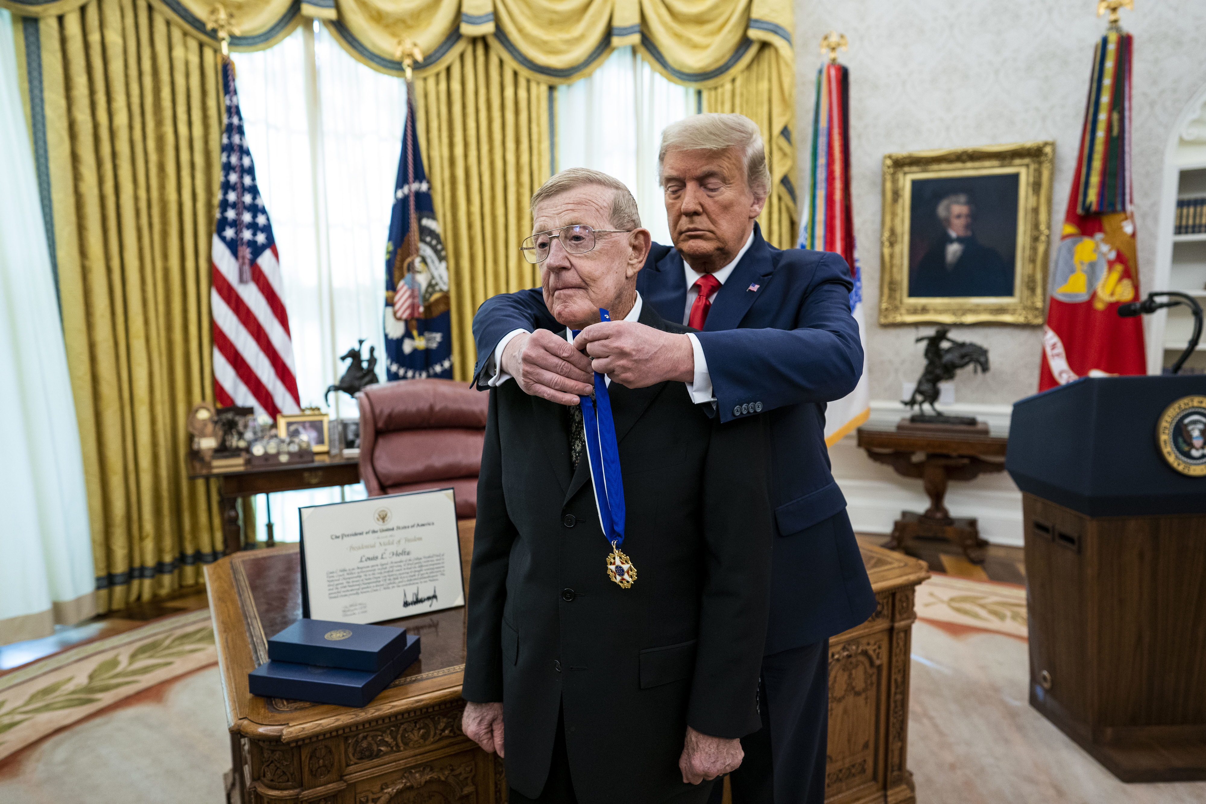 A man receives a medal from a person in a formal setting with a presidential seal on the desk