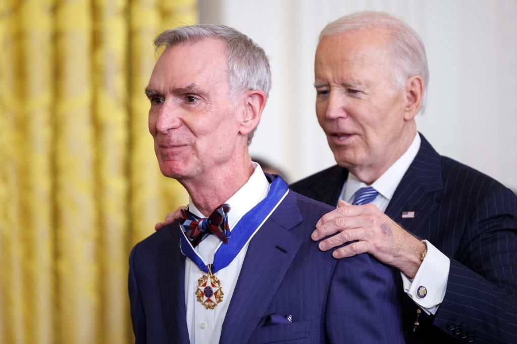 Two men at an award ceremony; one places a medal on the other, who wears a suit and bow tie