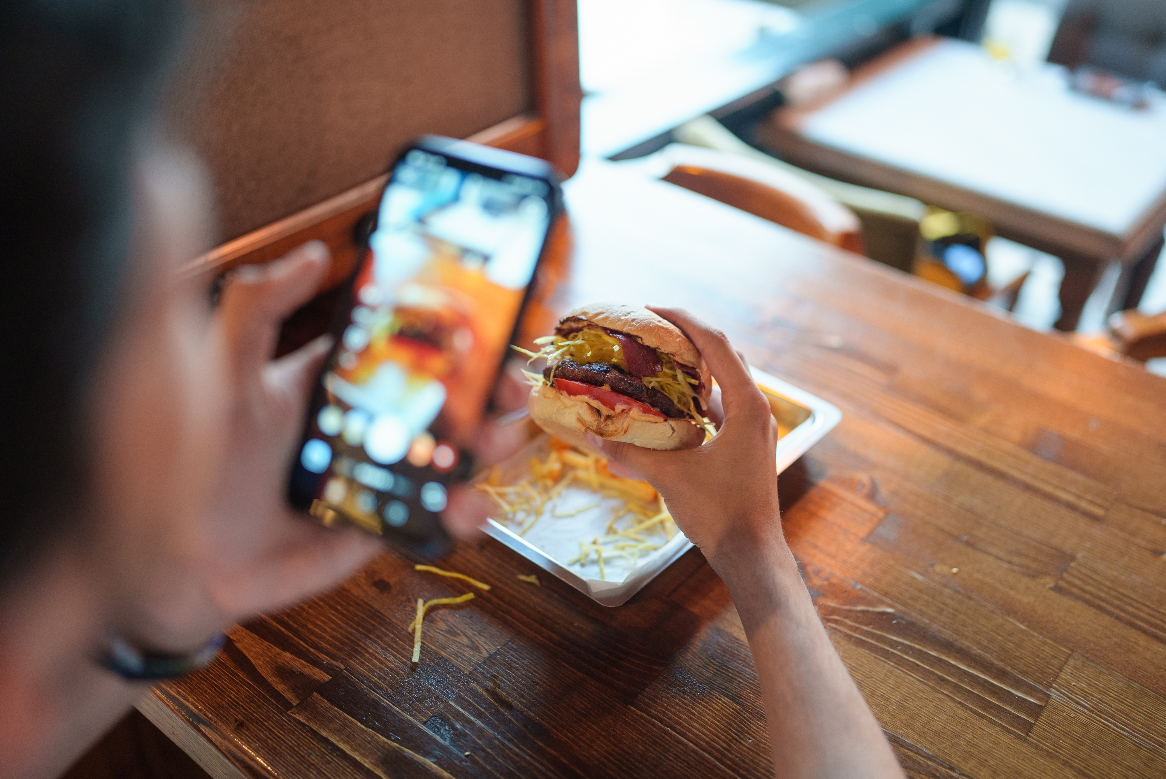 Person photographing a gourmet burger with a smartphone at a wooden table, capturing culinary details for social media