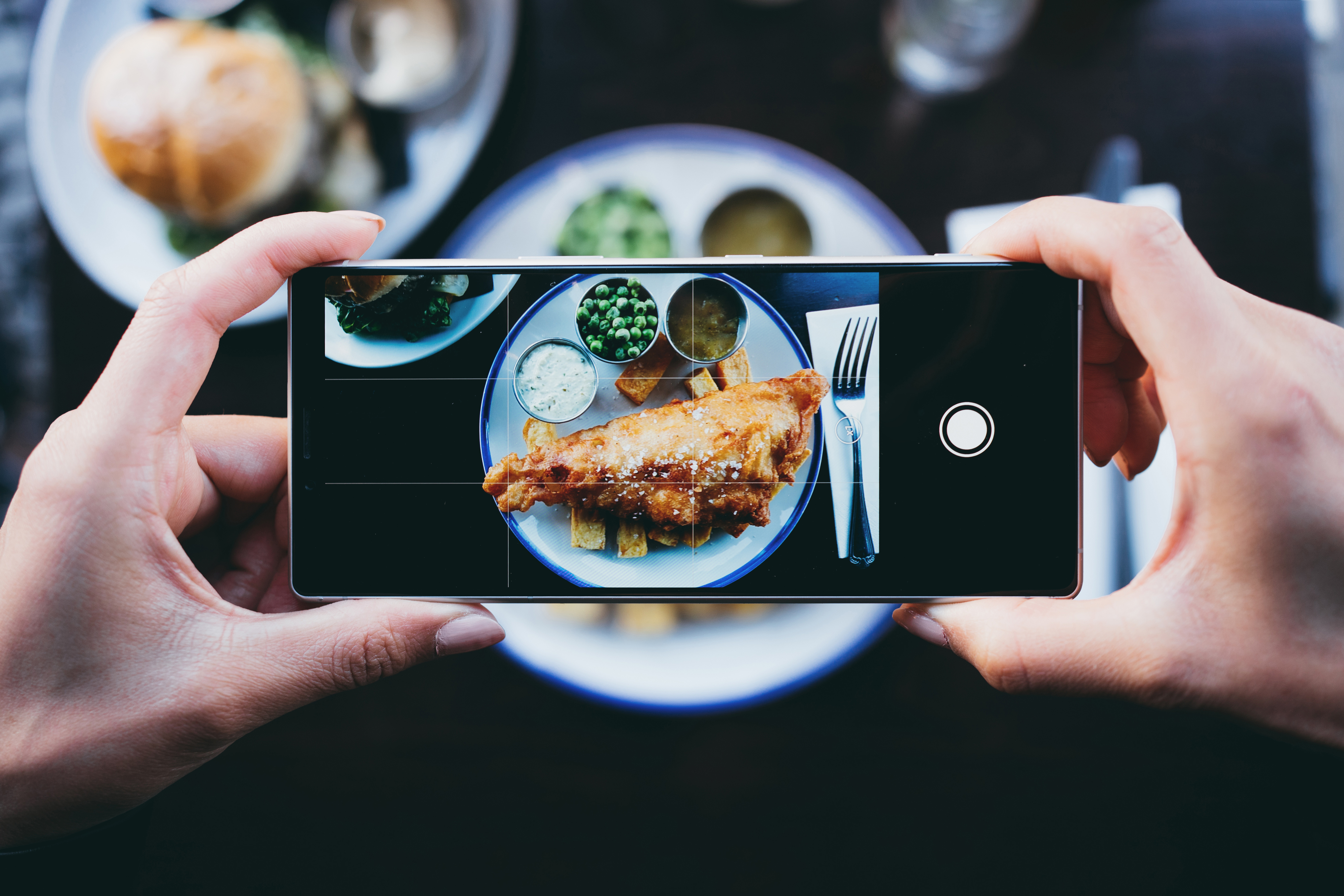 Person photographing a meal of fish and chips with peas and sauce