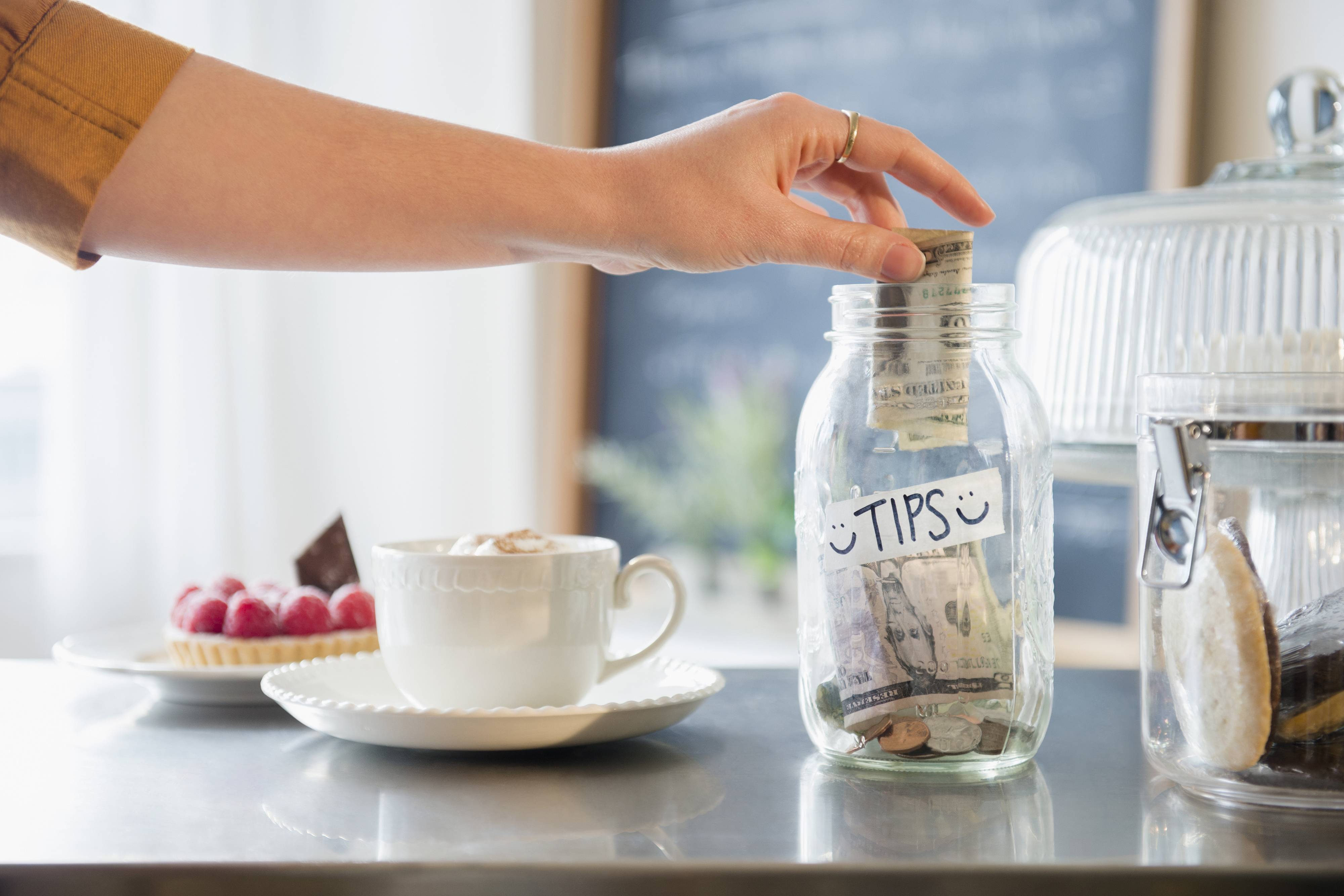 Hand placing cash in a tip jar labeled &quot;Tips&quot; beside a cappuccino and raspberry tart on a café counter