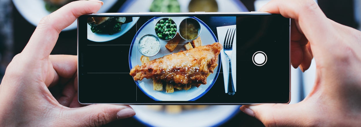 Person photographing fish and chips with peas on a plate at a restaurant table, blurred burger in the background