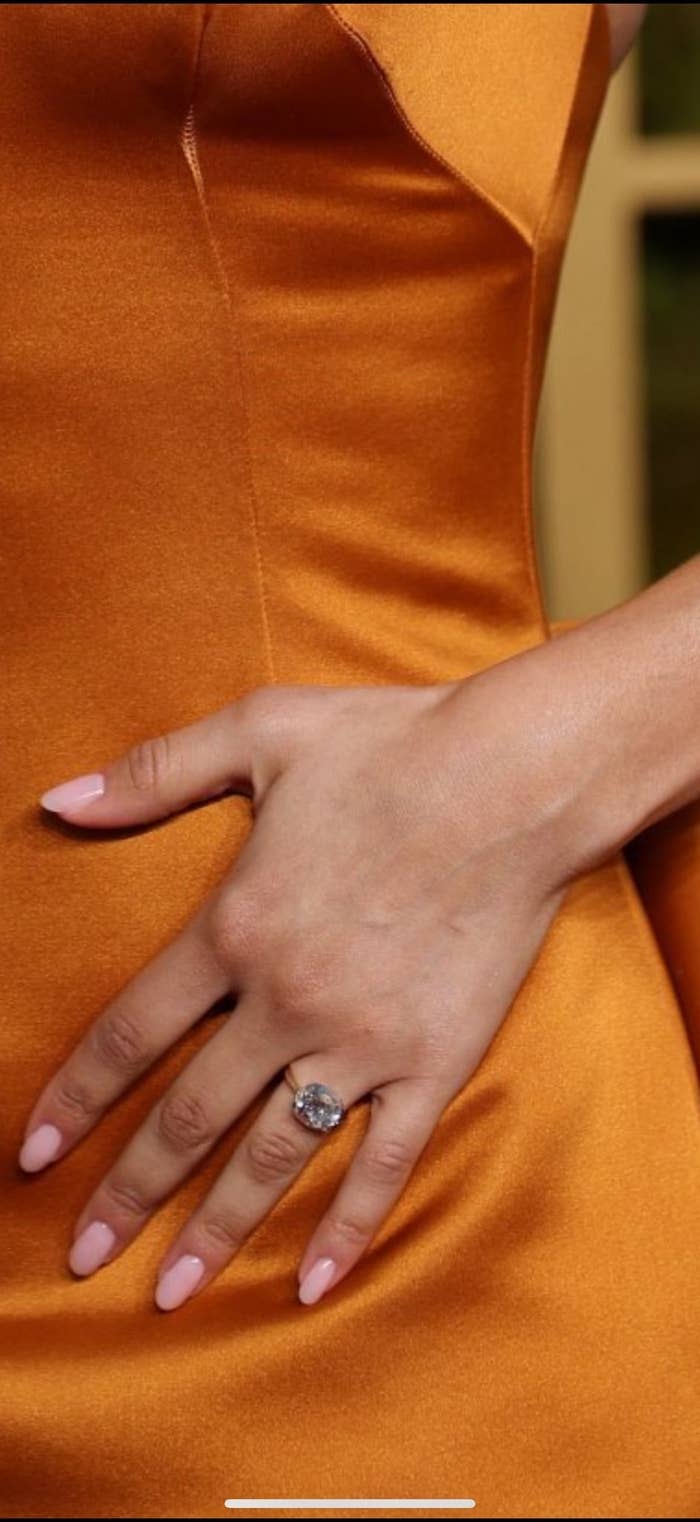 Primer plano de una mano con anillo grande y uñas cuidadas, posando sobre un vestido elegante
