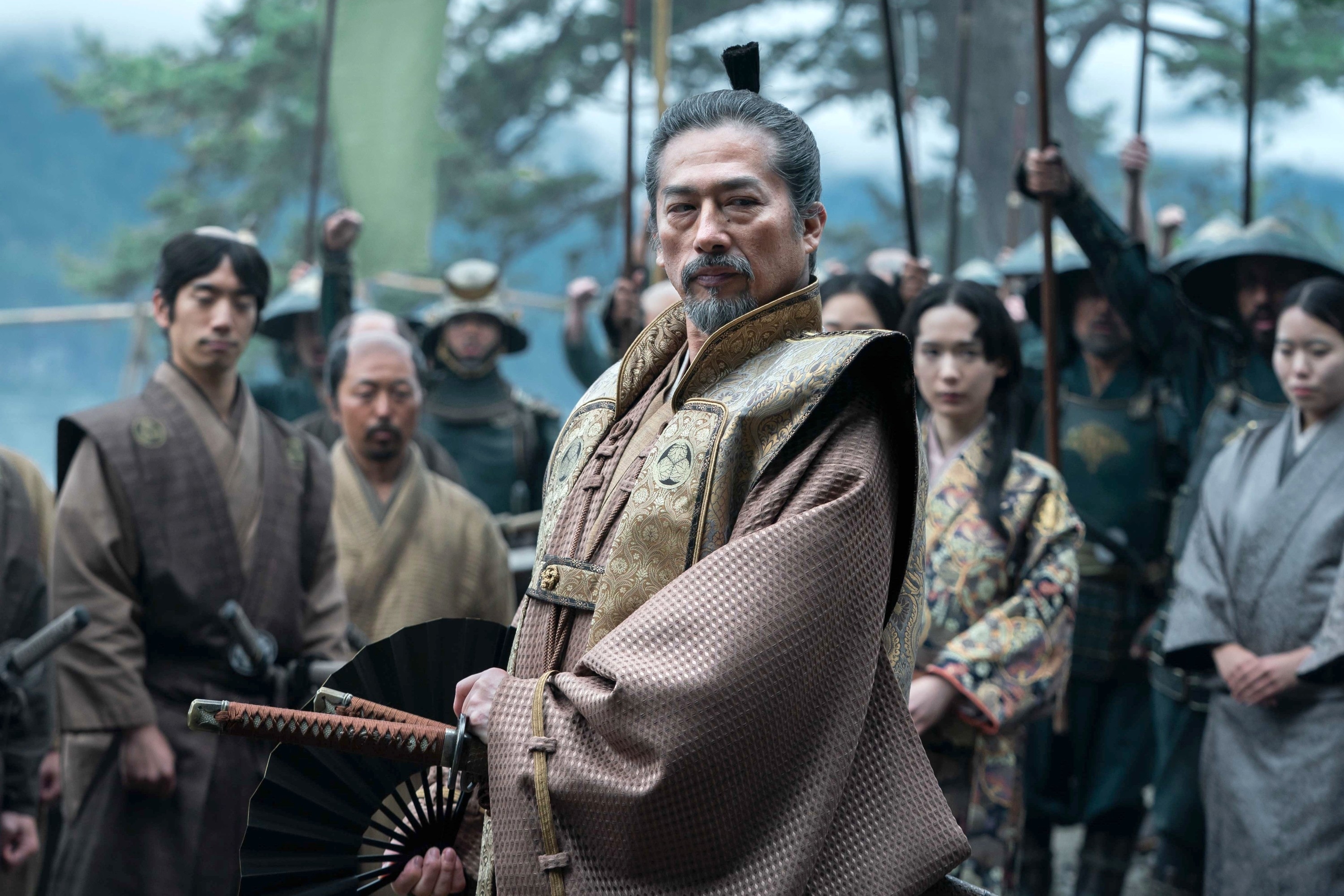 Scene from a historical drama with an older man in traditional clothing holding a fan, surrounded by others in period attire