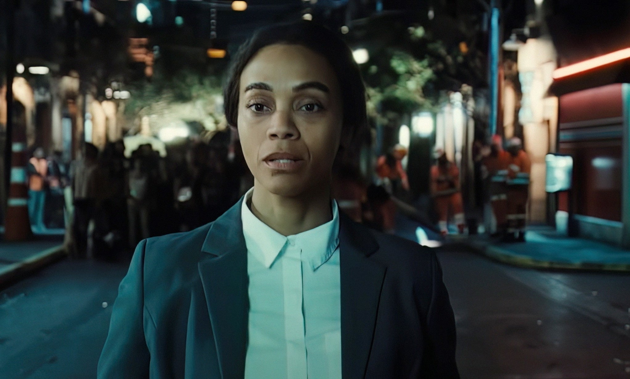 Person in a suit walking on a city street at night, looking determined. Background shows a small crowd and an illuminated building