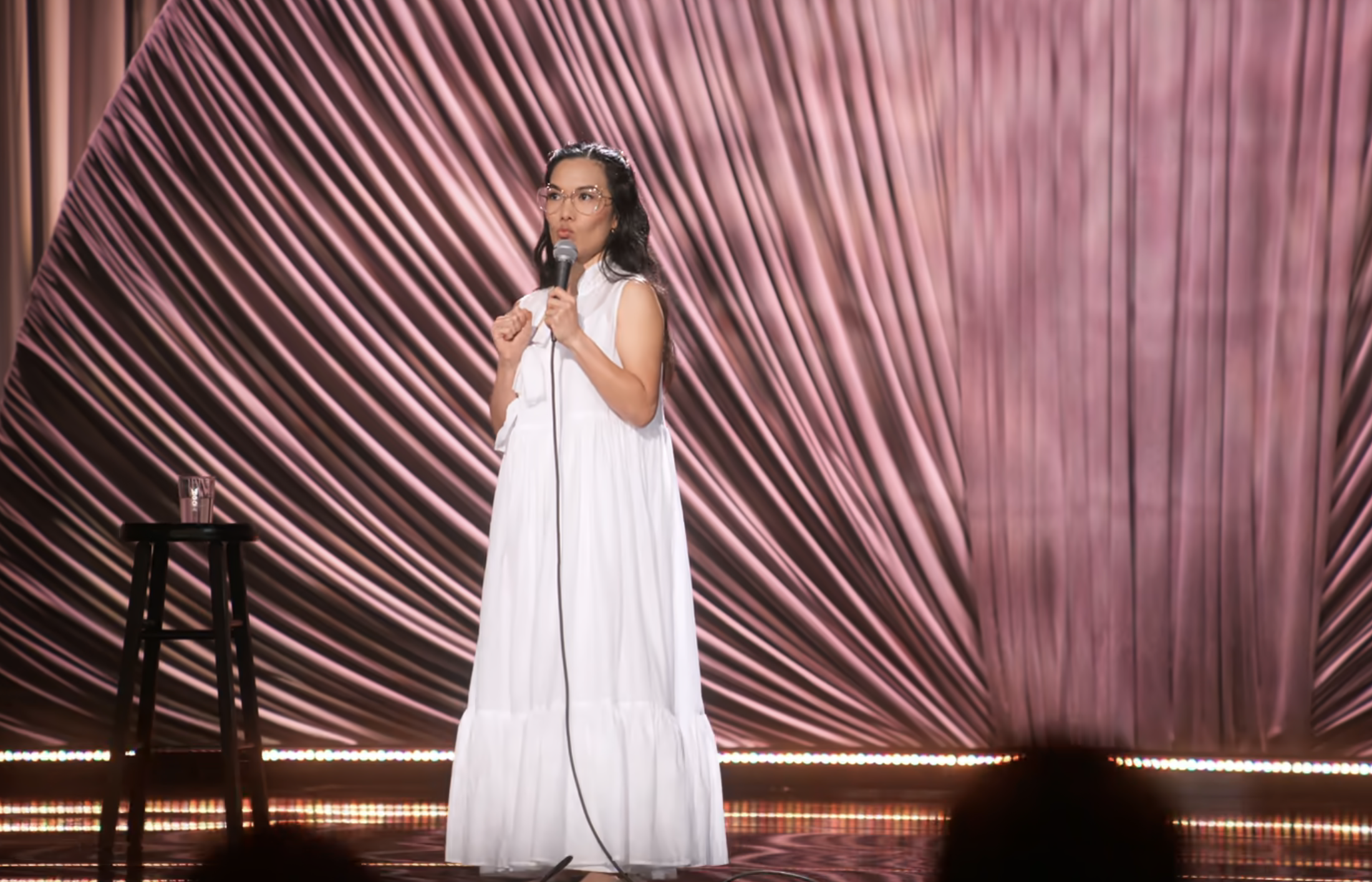 Comedian on stage with a microphone, wearing a flowy white dress, performing stand-up comedy