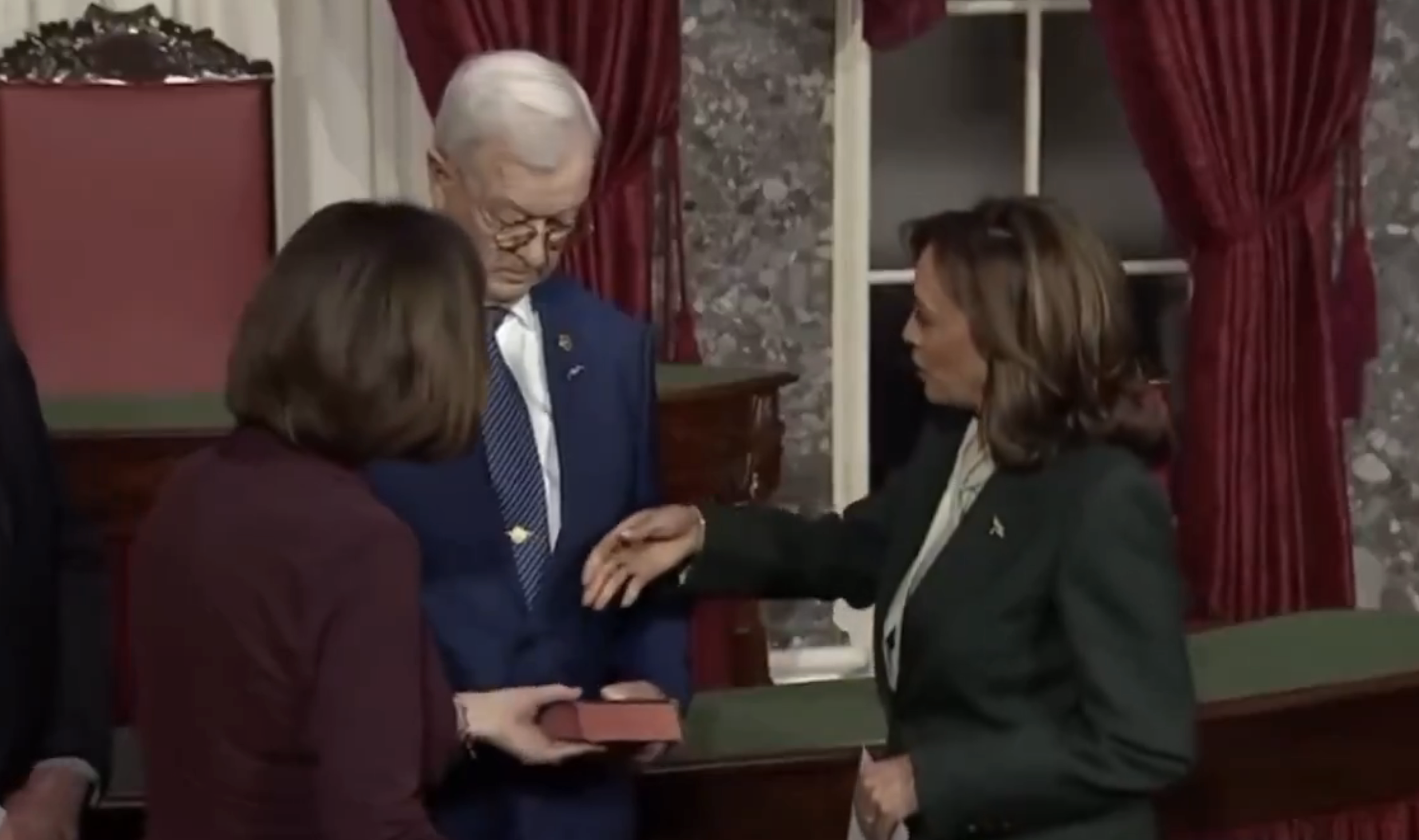 Three people at an official ceremony with one holding a book and another extending a right hand, possibly taking an oath