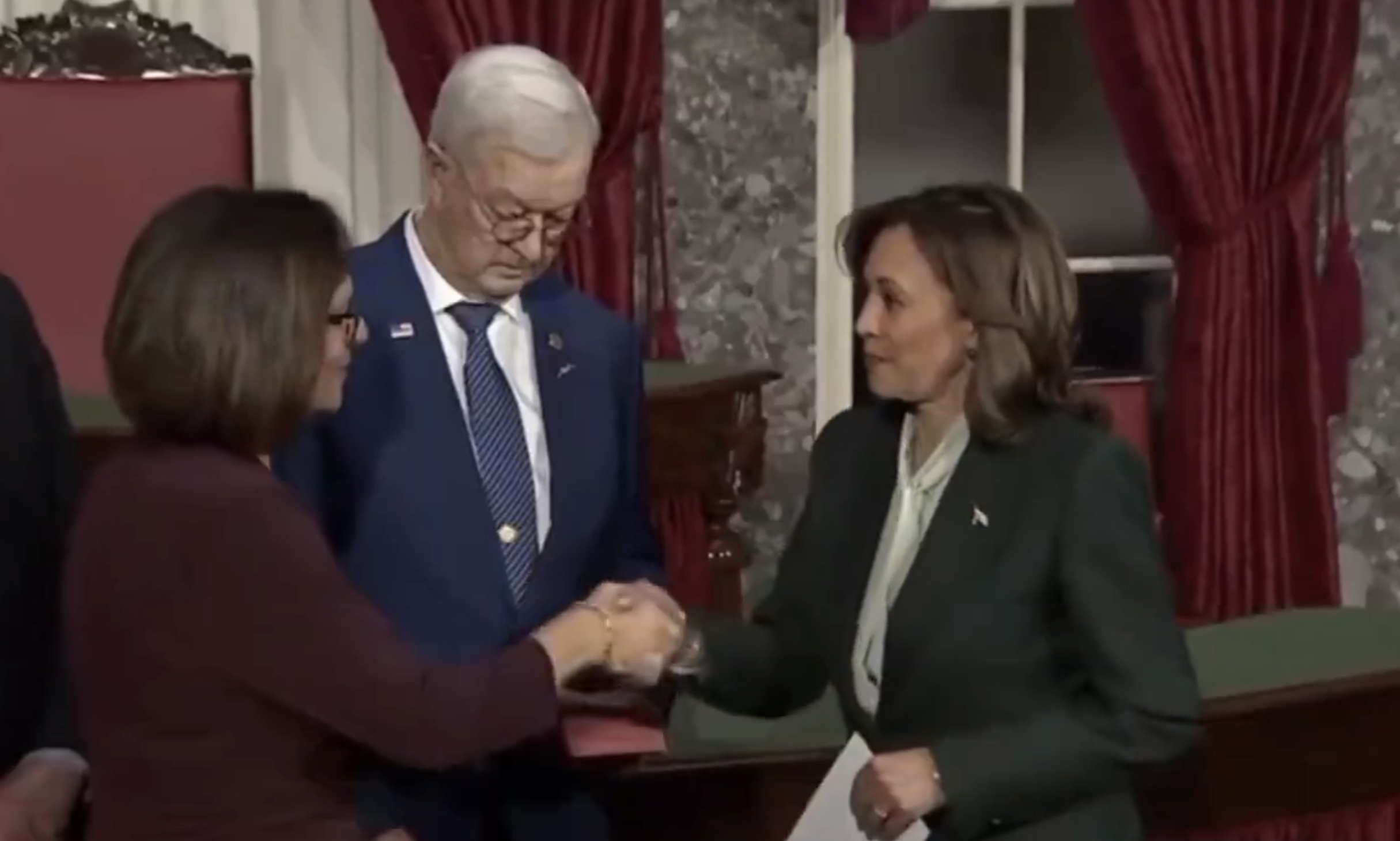 A woman in formal attire shakes hands with another woman in a suit, in a ceremonial setting with curtains and marble walls in the background