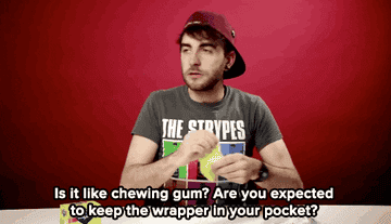 Man with cap on backwards opens a gum pack, reads text: &quot;Is it like chewing gum? Are you expected to keep the wrapper in your pocket?&quot;