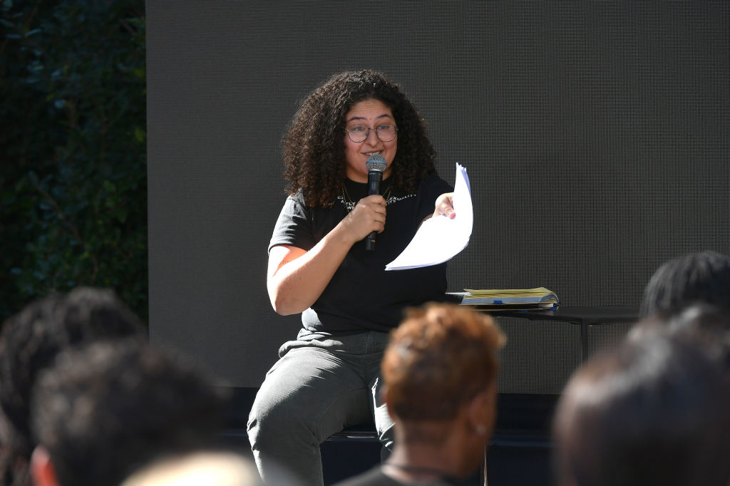 Person with curly hair and glasses sits, holding a microphone and papers, speaking to an audience seated outdoors