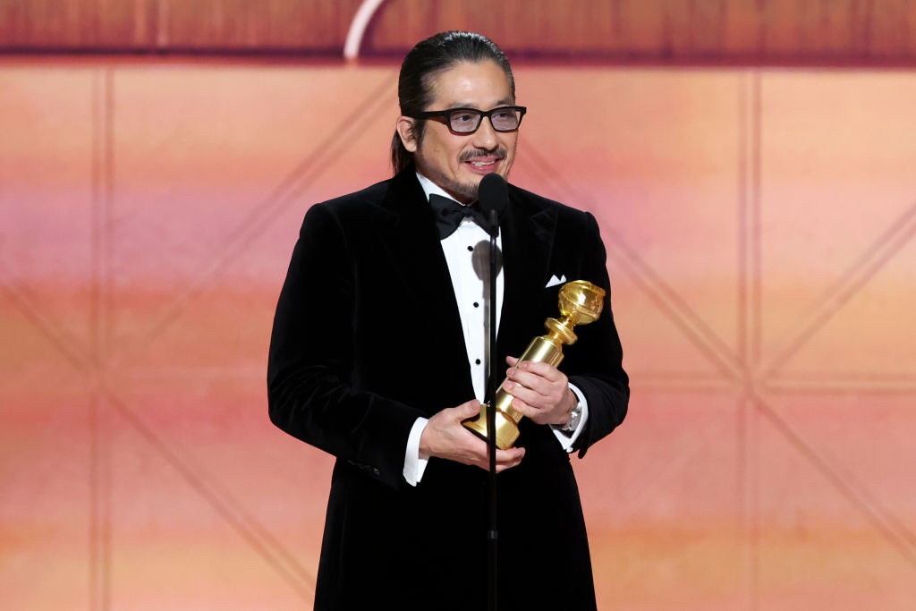 Hiroyuki Sanada in a tuxedo holds a trophy on stage at an awards ceremony, speaking into a microphone