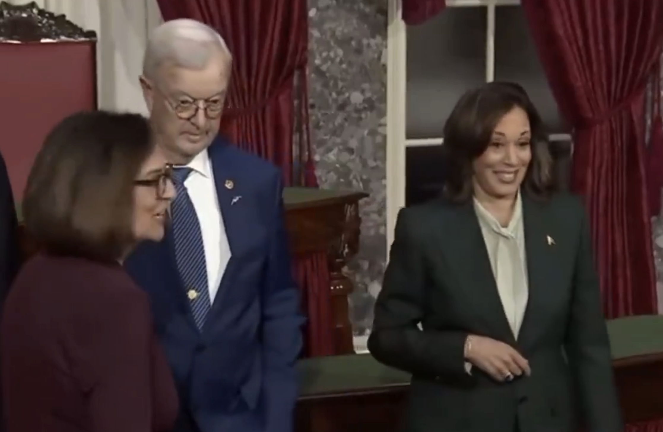 Three people are standing together. A woman in a suit is smiling next to a man dressed formally. The setting suggests a formal or official event