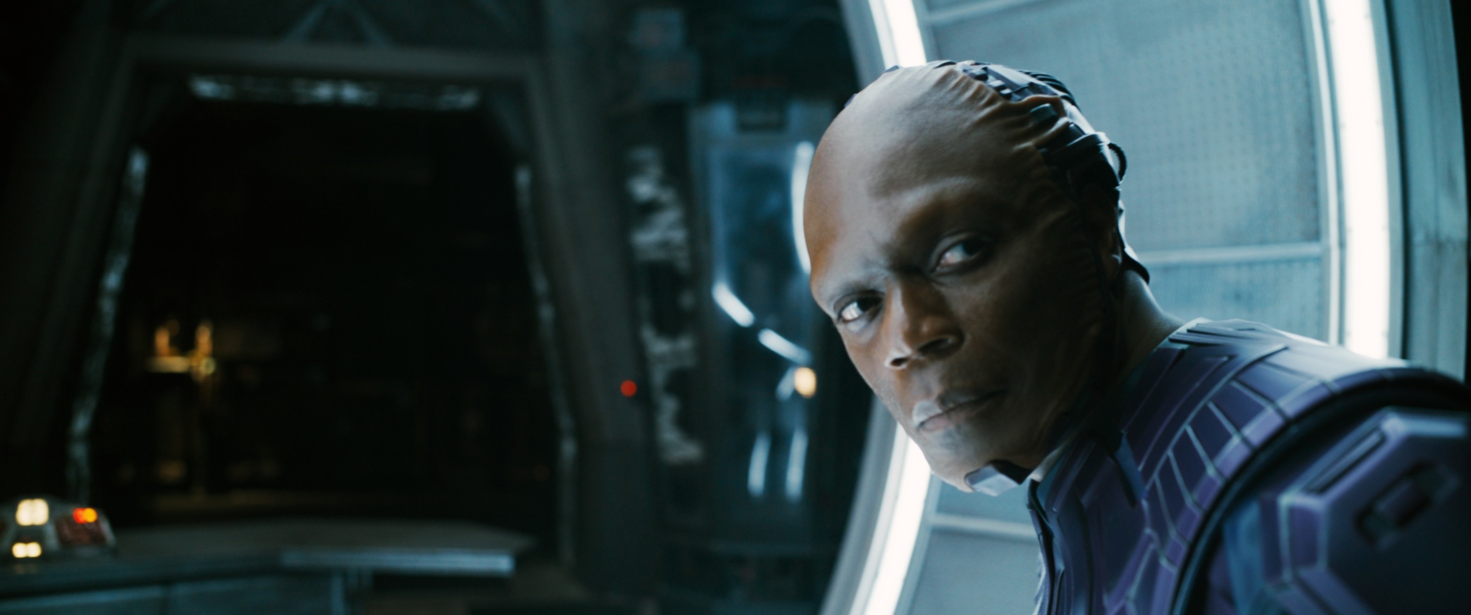 Alien character in a futuristic setting stands in a spaceship, wearing geometric-patterned armor, looking towards the camera